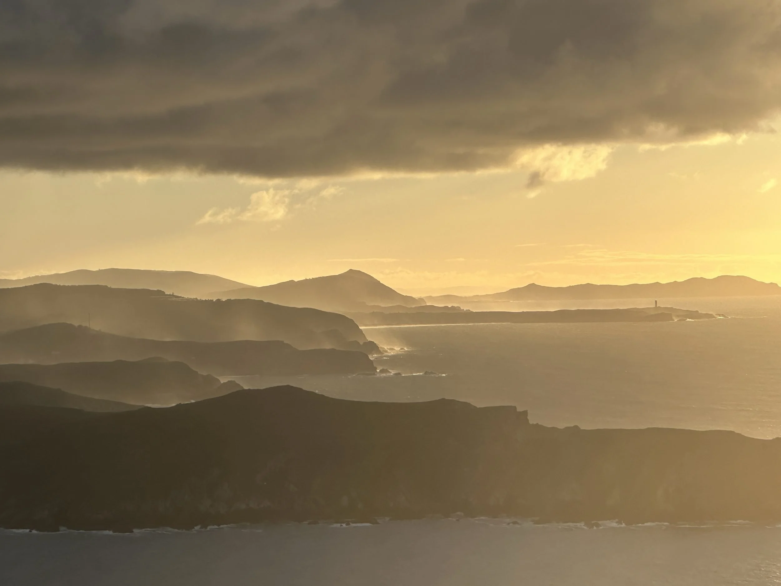 Wide-angle landscape of layered coastal mountain silhouettes veiled in golden evening haze and soft sea mist along the Northern Galician coastline, photographed by Seña Studio.