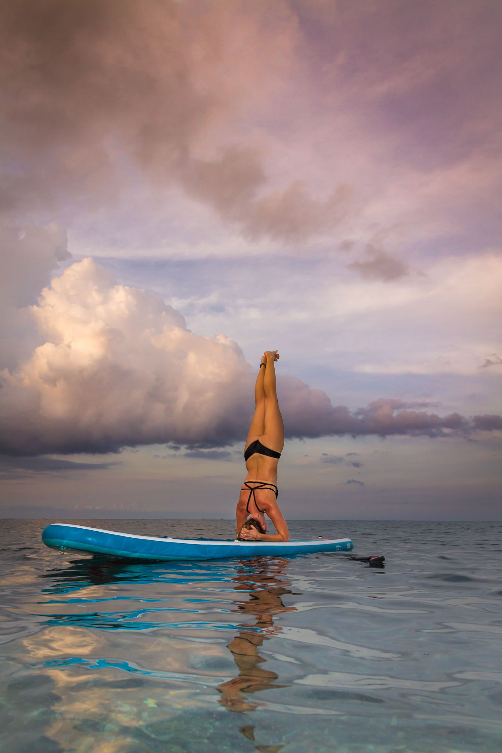 Full-length shot of a woman performing a vertical headstand yoga pose on a paddleboard over calm, reflective ocean water under a cloudy sunset sky, photographed by Seña Studio.