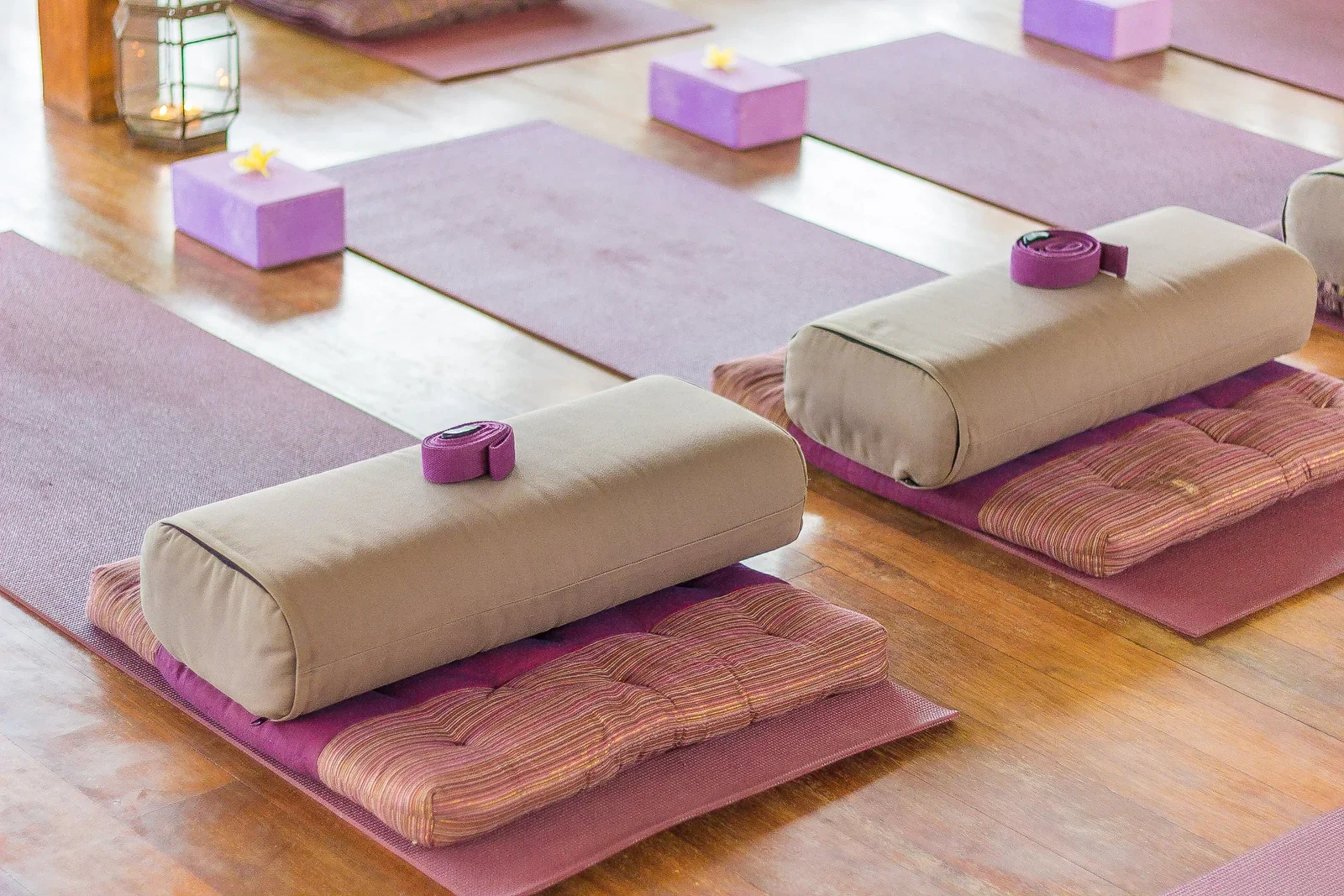 Organized yoga and meditation room arrangement on polished hardwood floors, featuring purple mats, beige bolsters, rolled straps, and purple foam blocks topped with frangipani blossoms.
