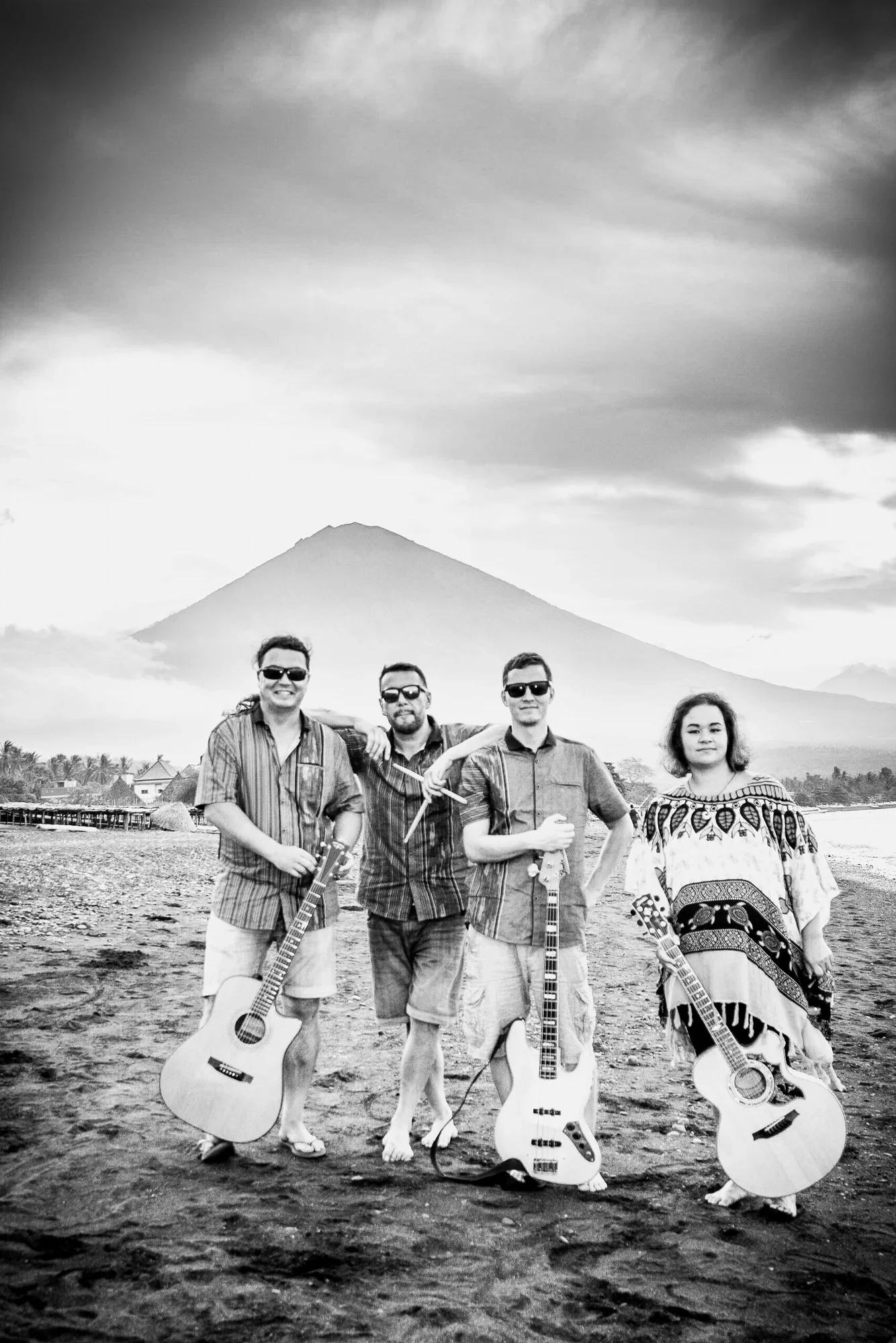 Black-and-white environmental band portrait on a sandy beach, posing with acoustic guitars and bass, framed by Mount Agung in the background, photographed by Seña Studio. Musician Lucy Dobrovodska leads the band.