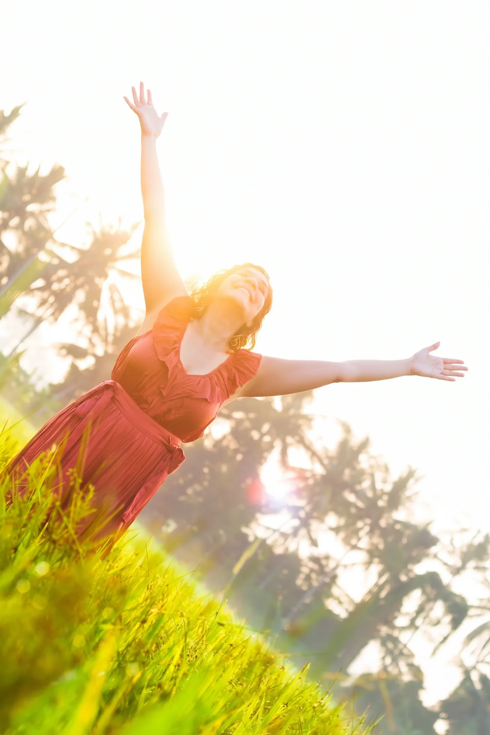 Low-angle shot of a woman in a terracotta dress joyfully raising her hands toward a warm golden sunflare over palm trees and green fields, photographed by Seña Studio.
