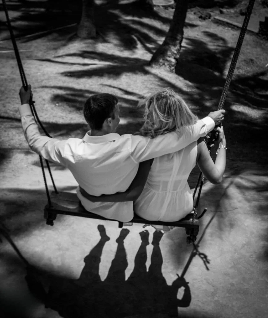 High-angle black-and-white portrait of a couple embracing on a large wooden rope swing, casting long shadows onto the ground below under a tropical canopy, photographed by Seña Studio.