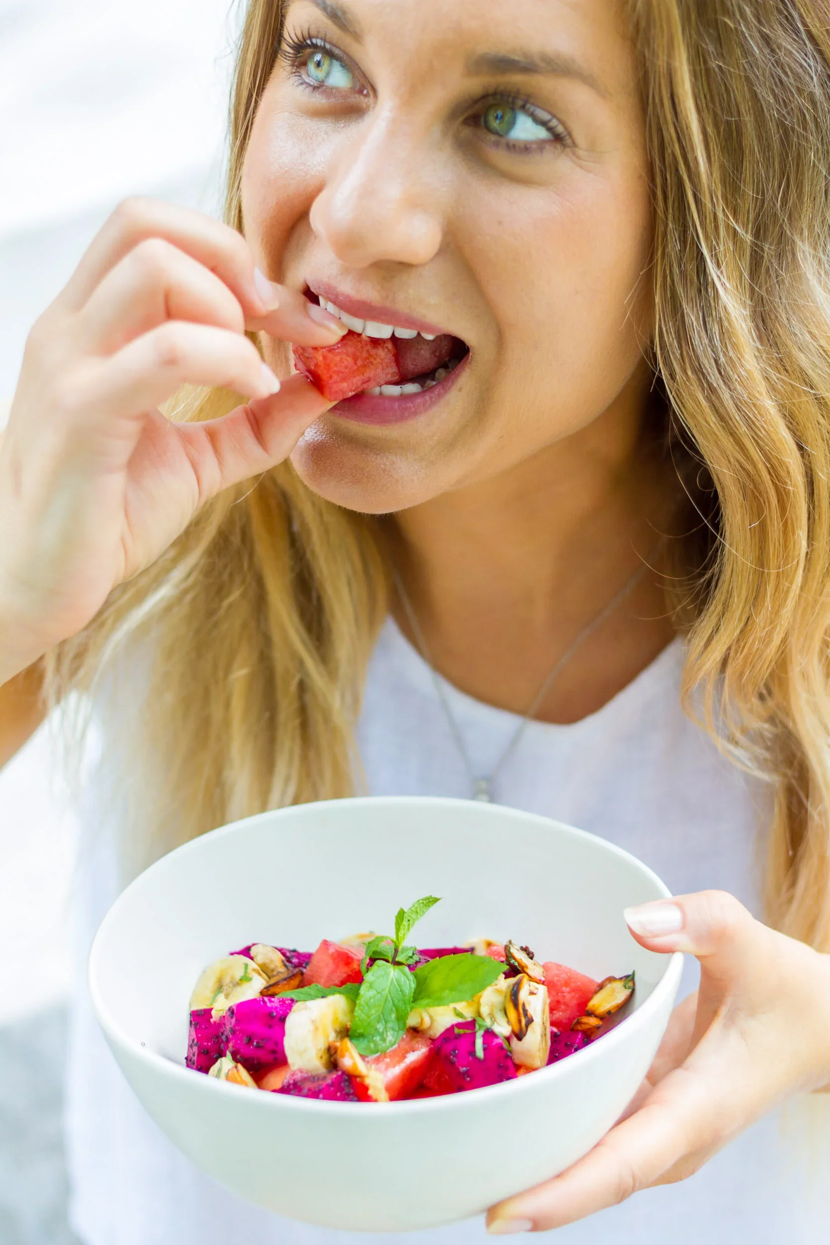 A close-up view of a smiling blonde woman eating a cube of watermelon while holding a white bowl filled with fresh dragon fruit, bananas, and mint.