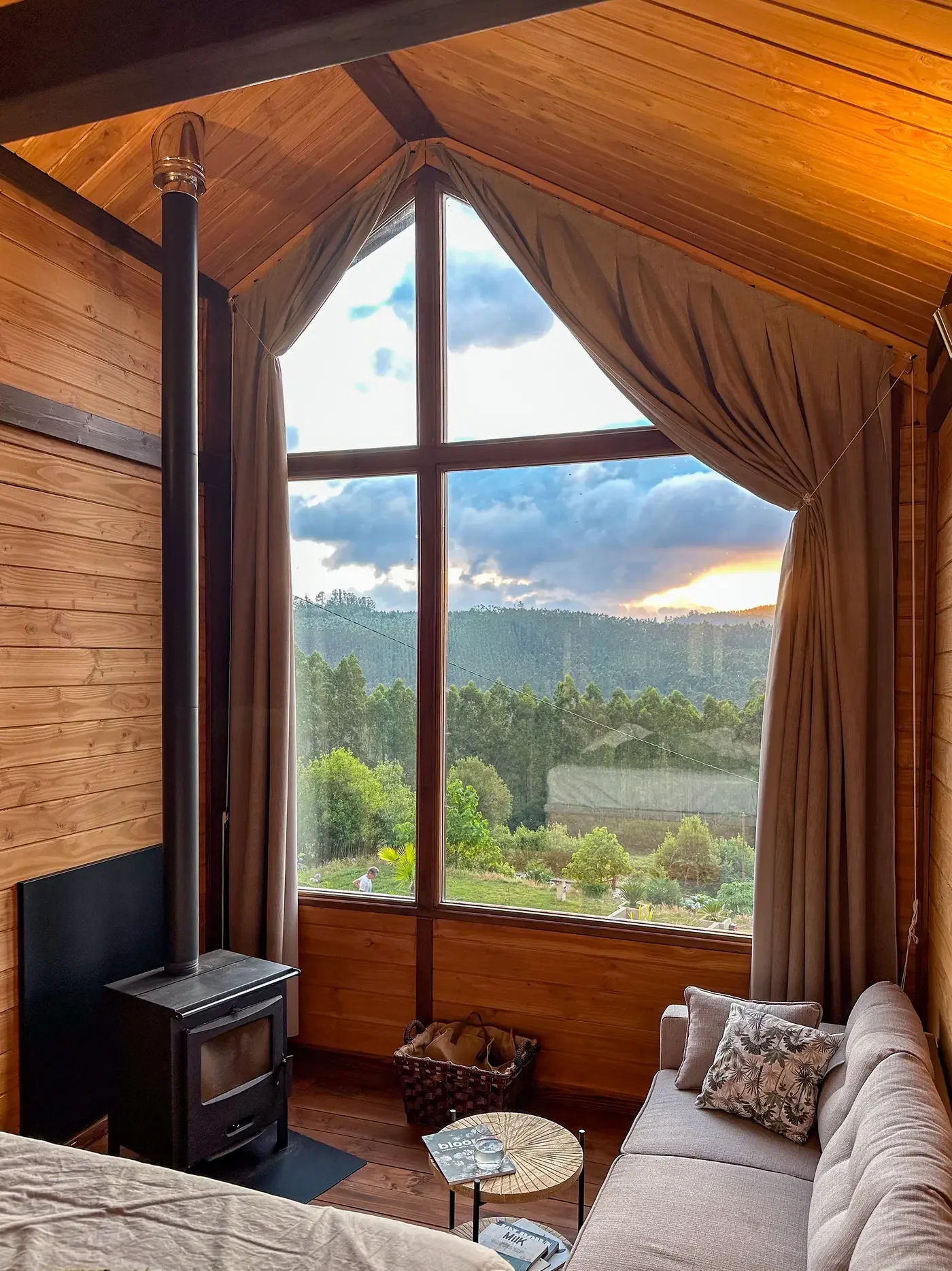 Cozy cabin interior featuring a large floor-to-ceiling A-frame window, wood-burning stove, and sofa overlooking scenic forest hills at Haras de Bordo, photographed by Seña Studio.
