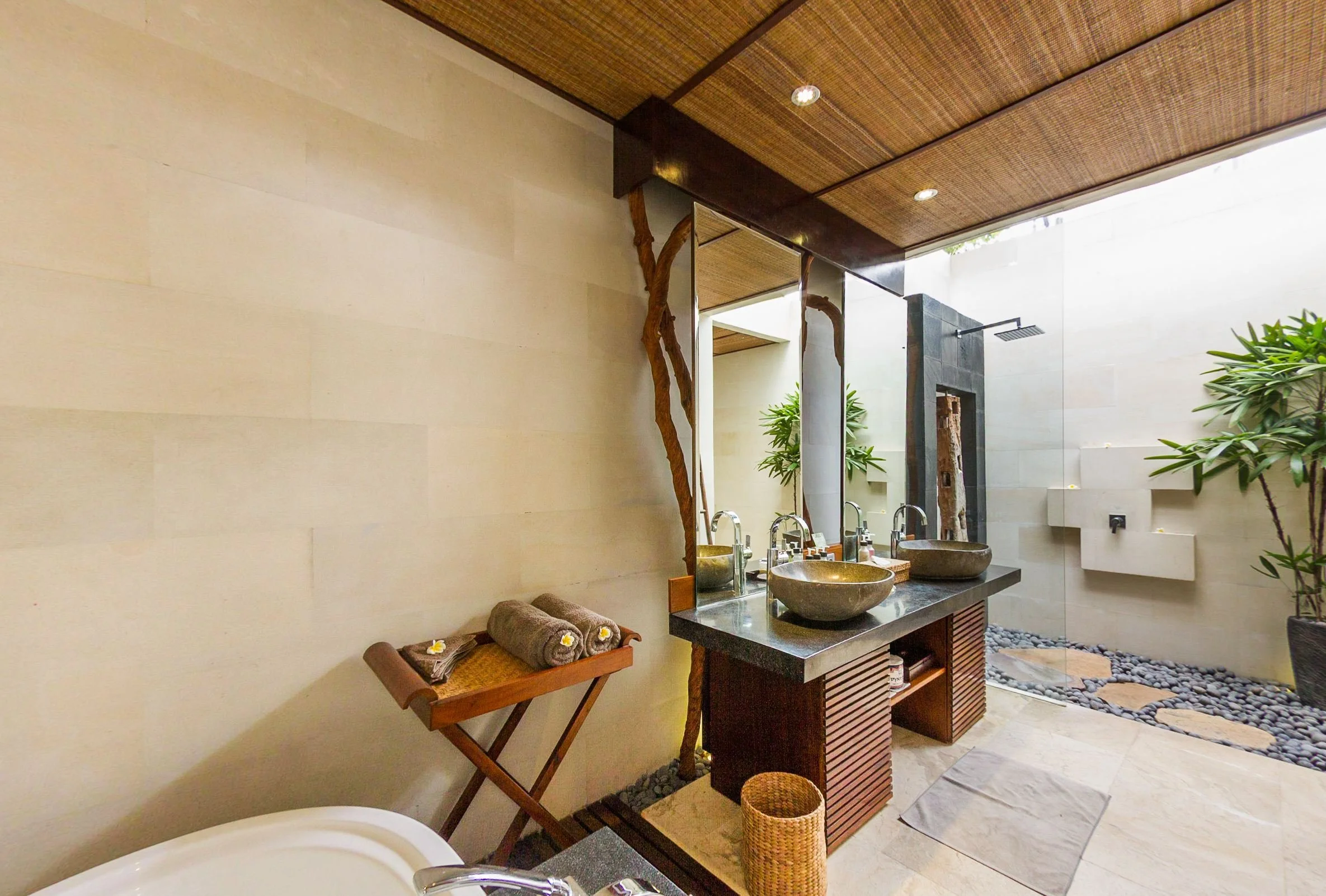 Spas-style open-air villa bathroom with stone vessel sinks, dark wood vanity, woven ceiling details, and tropical indoor plants at The Griya Villas & Spa in Amed, Bali, photographed by Seña Studio.
