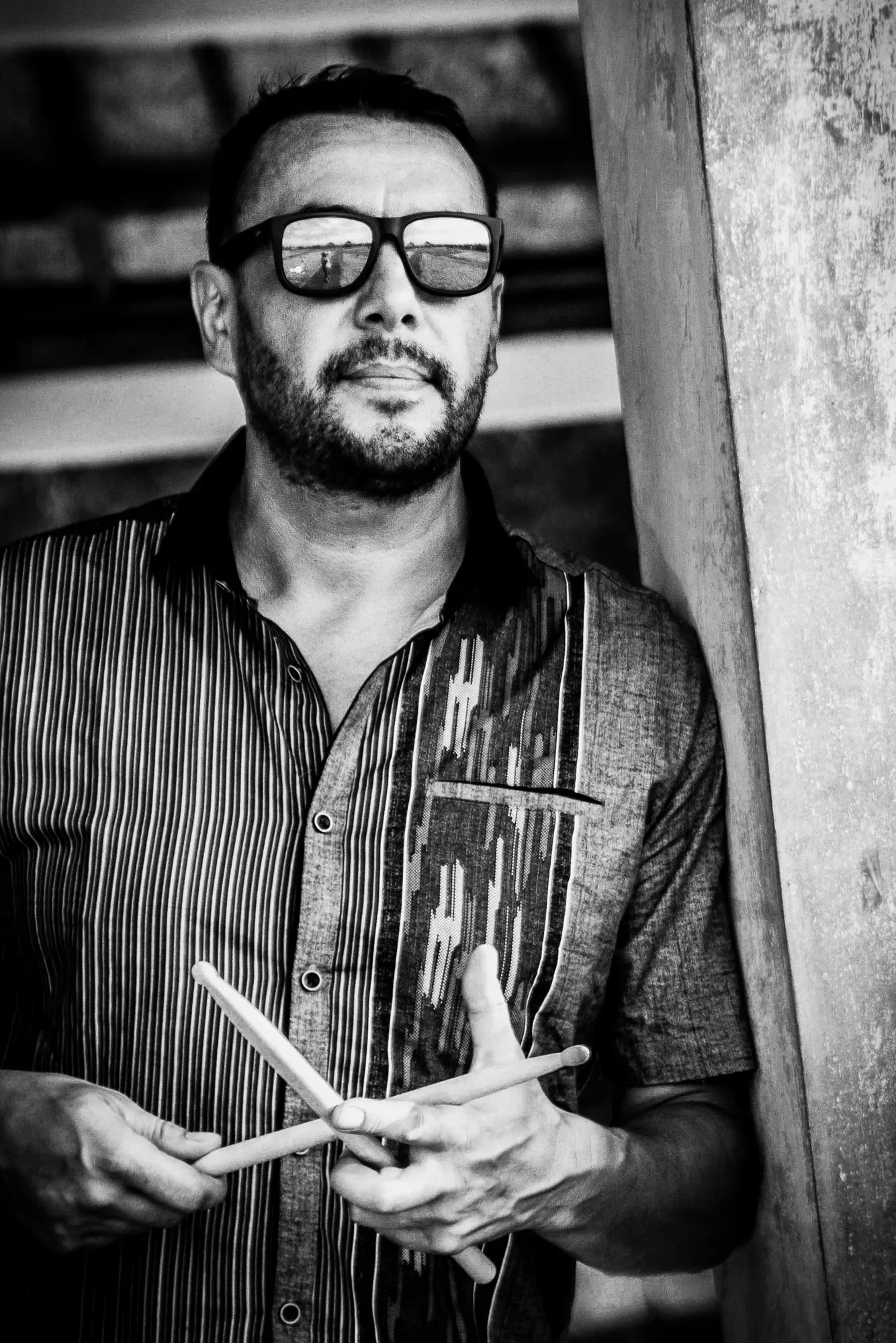 High-contrast black-and-white portrait of a male drummer holding crossed drumsticks, wearing reflective sunglasses that capture the beach horizon, photographed by Seña Studio.