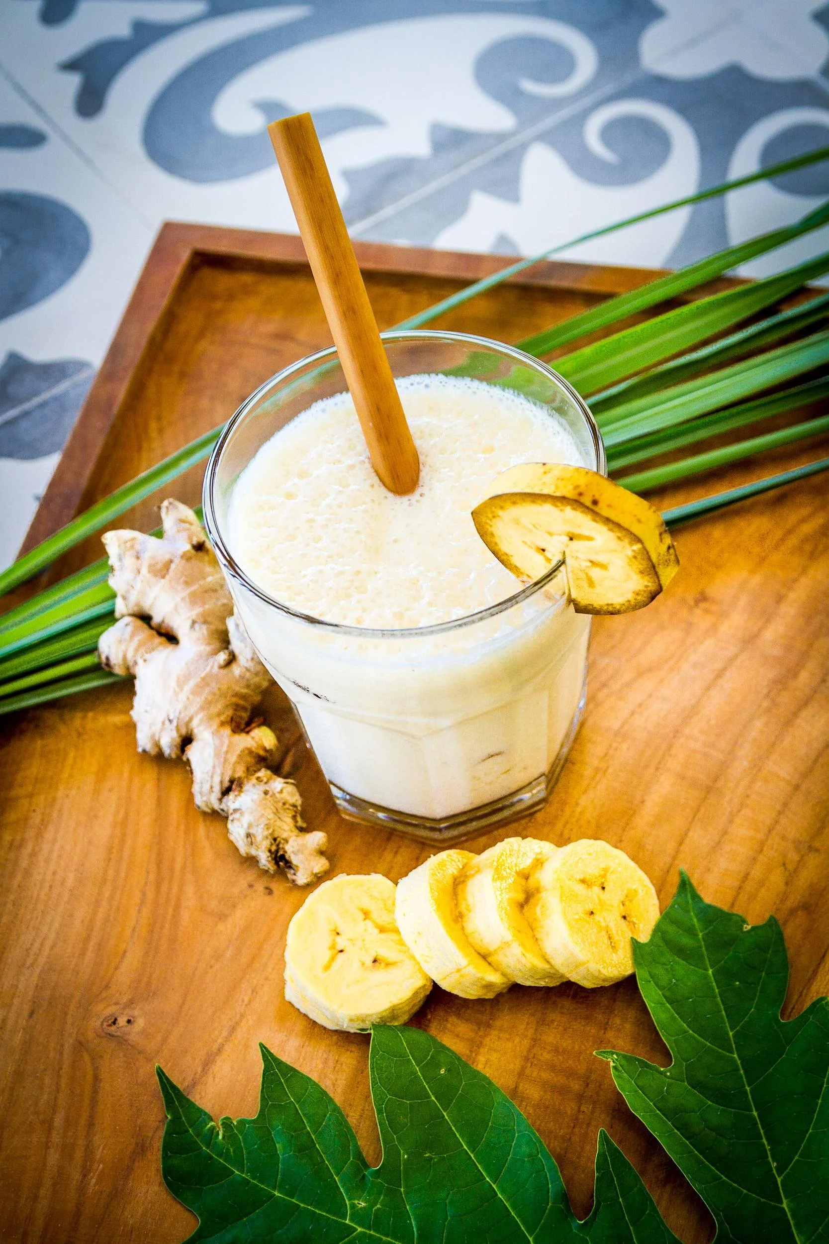 High-angle photography of a banana ginger smoothie in a glass with a bamboo straw, surrounded by sliced bananas, fresh ginger root, and green leaves on a wooden tray, photographed by Seña Studio.