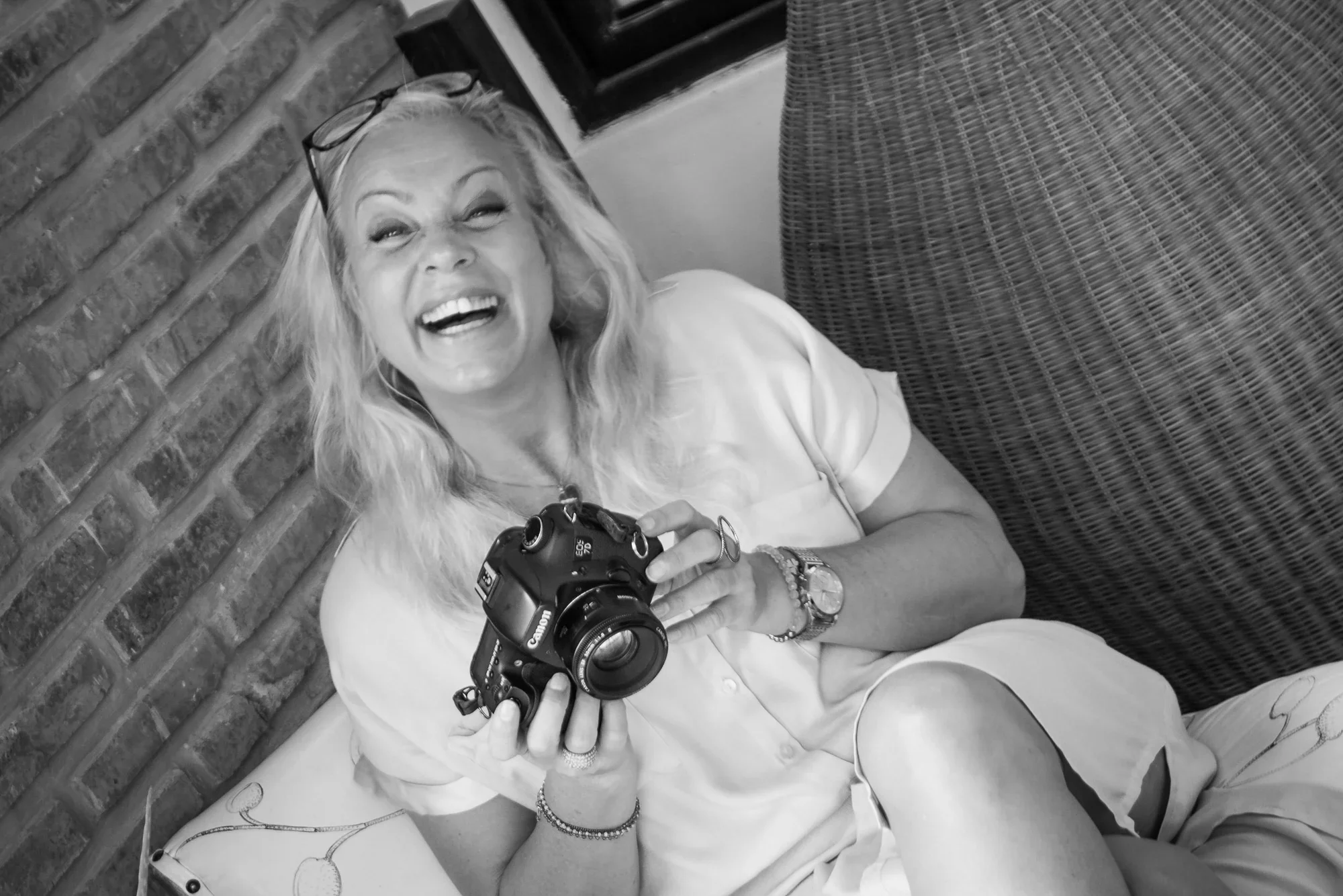 Behind-the-scenes, black-and-white photograph of a laughing blonde woman holding a black Canon DSLR camera during a photoshoot.