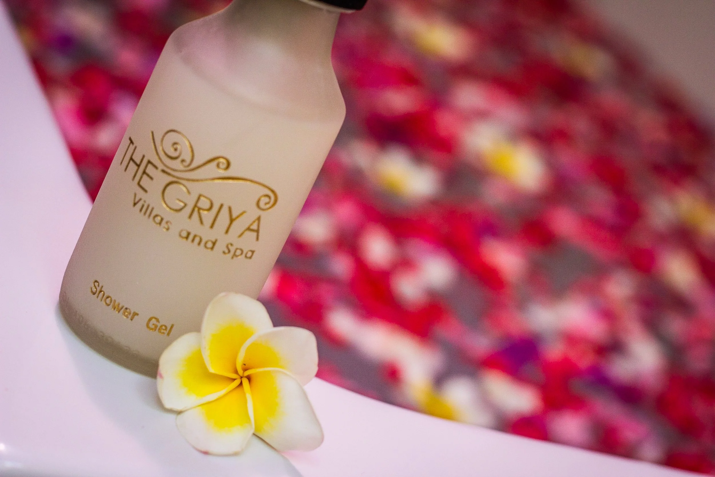 Close-up of a branded luxury shower gel bottle and a yellow frangipani flower resting on the edge of a bathtub filled with red flower petals at The Griya Villas & Spa, photographed by Seña Studio.