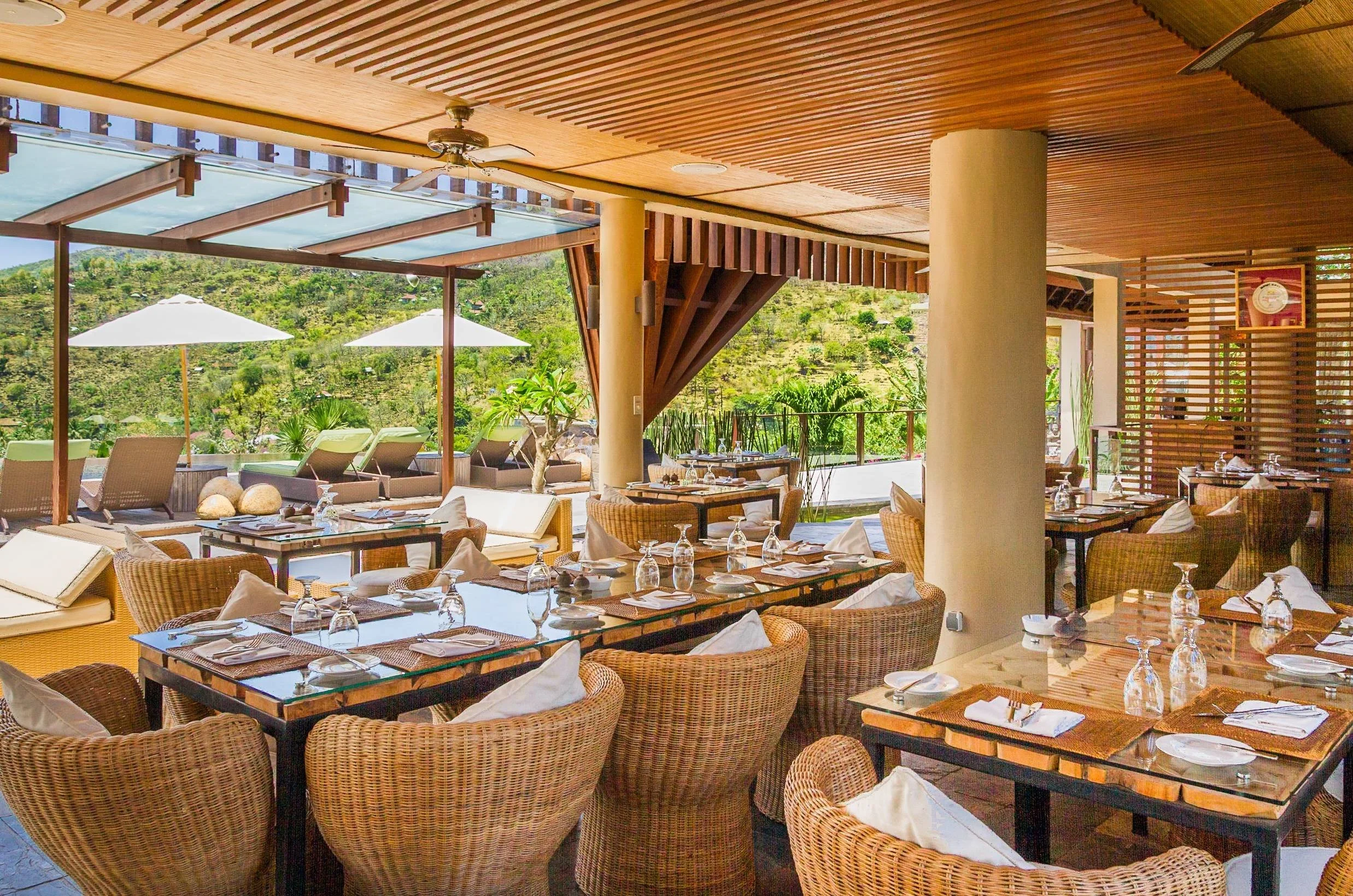 Open-air luxury resort restaurant interior with woven rattan dining chairs, set glass-top tables, and wooden slatted ceiling looking out toward green hills, photographed by Seña Studio.