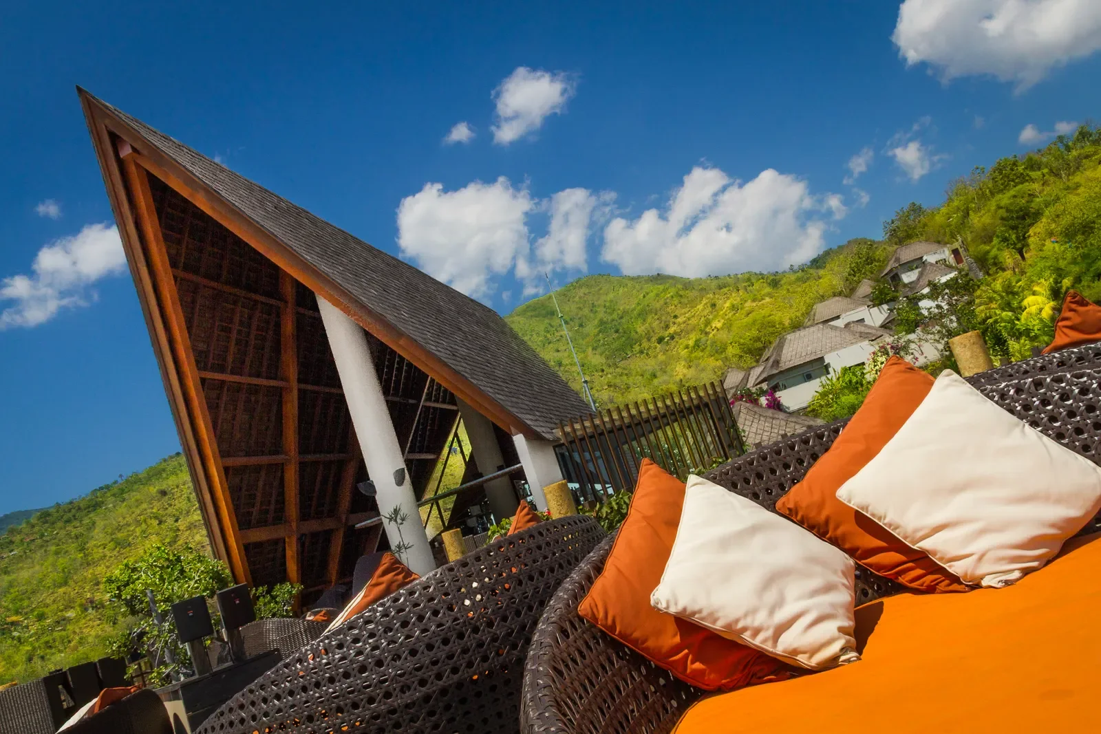 Wide-angle shot of outdoor lounge seating with orange cushions beneath a dramatic thatched pavilion against blue skies and mountain views at The Griya Villas & Spa in Amed, Bali, photographed by Seña Studio.