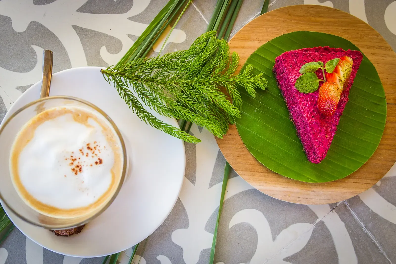 Top-down view of a frothy café latte and a vibrant pink dragon fruit cheesecake slice on a banana leaf at The Griya Villas & Spa in Amed, Bali, photographed by Seña Studio.