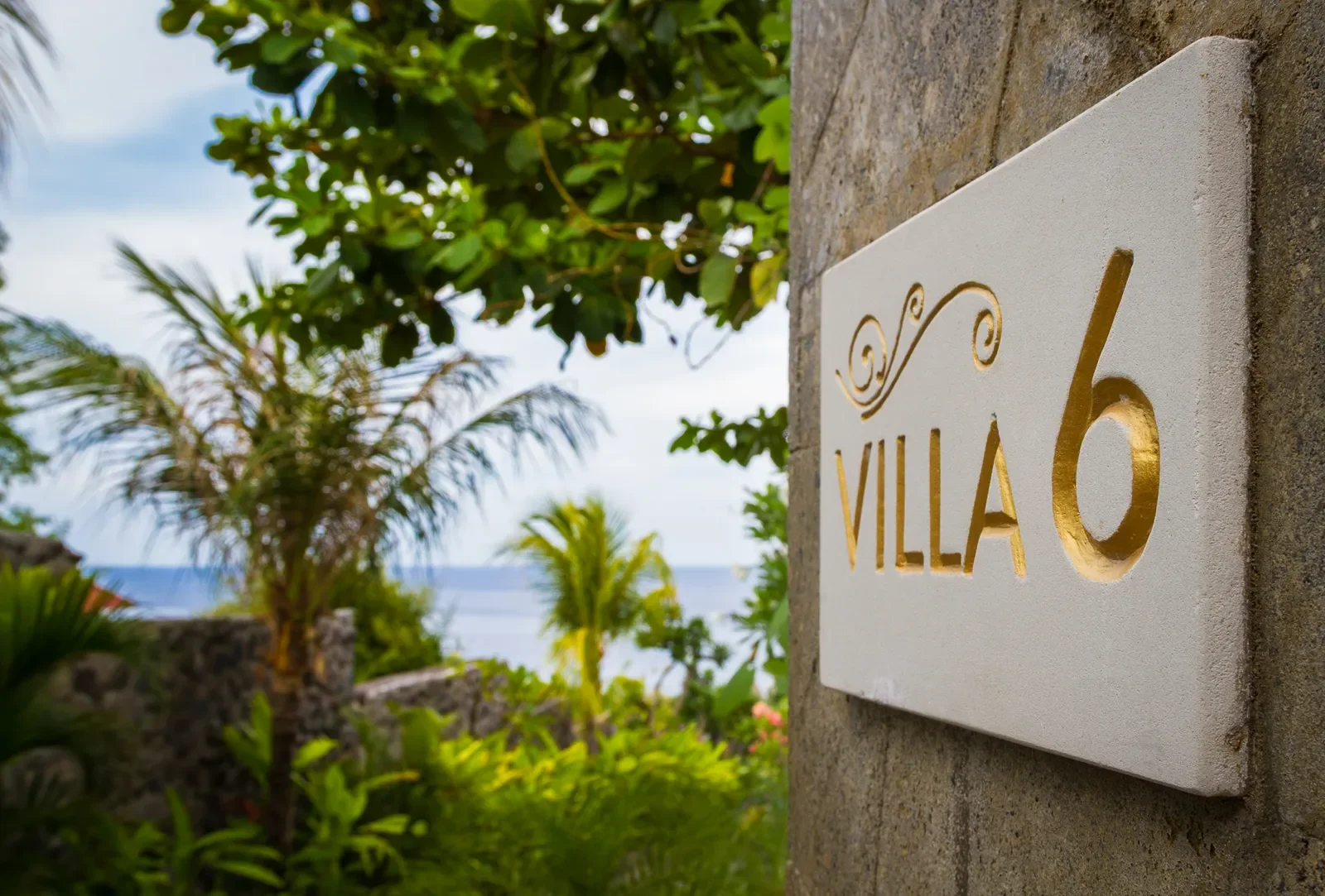 Engraved Villa 6 stone entrance sign surrounded by lush tropical greenery with an ocean view in Amed, Bali, photographed by Seña Studio.