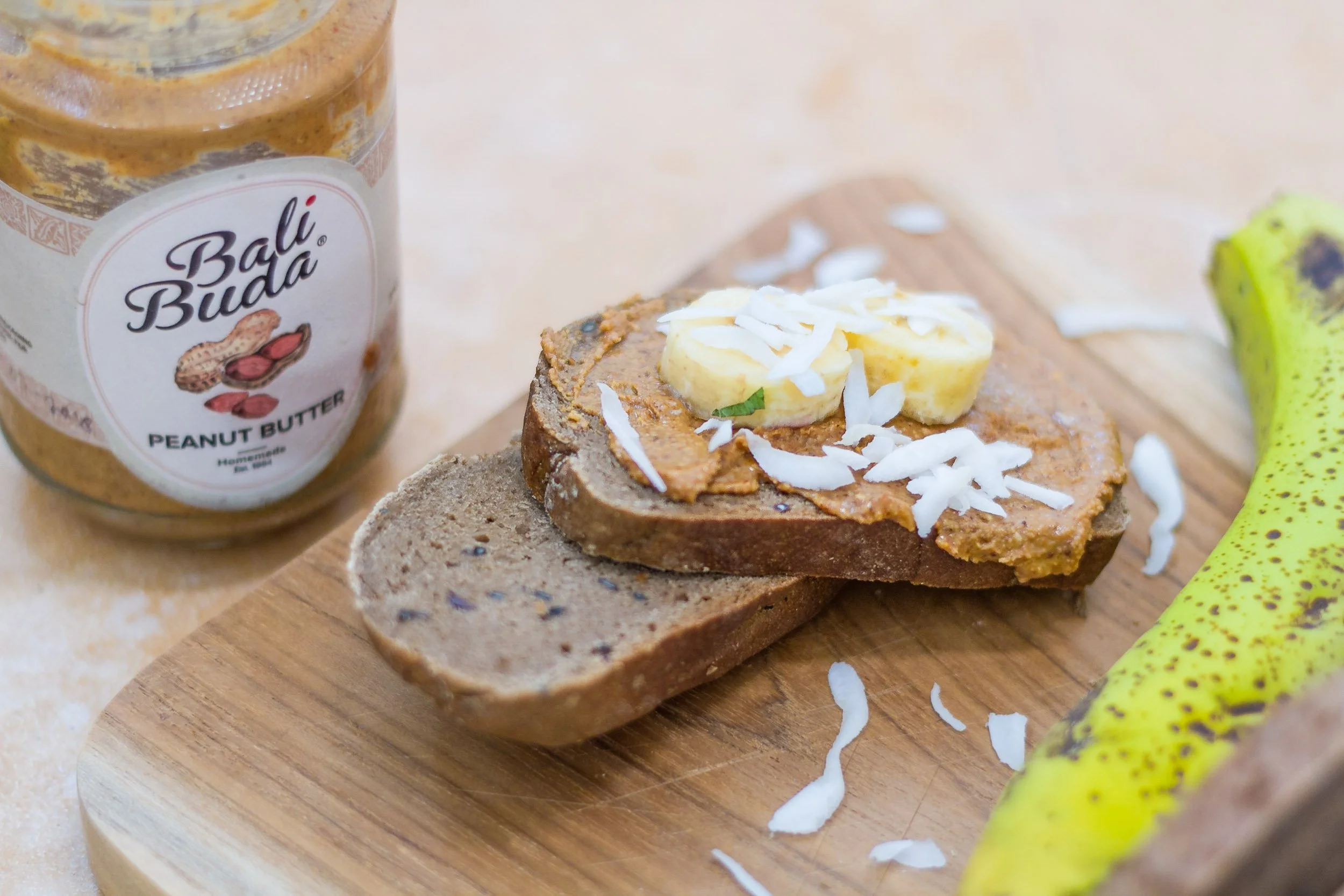 Sliced dark artisanal bread spread with Bali Buda peanut butter and topped with banana slices and shredded coconut on a wooden board next to a banana and peanut butter jar.