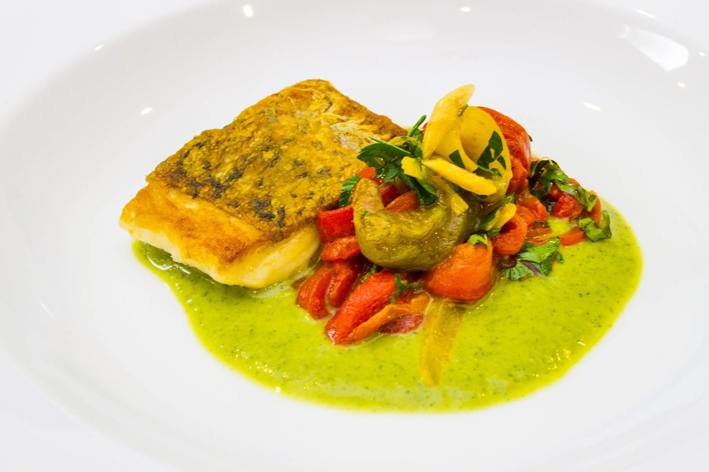 Plated fine-dining shot of a pan-seared white fish fillet with crispy skin, served on a vibrant green herb puree and garnished with roasted red peppers, photographed by Seña Studio.