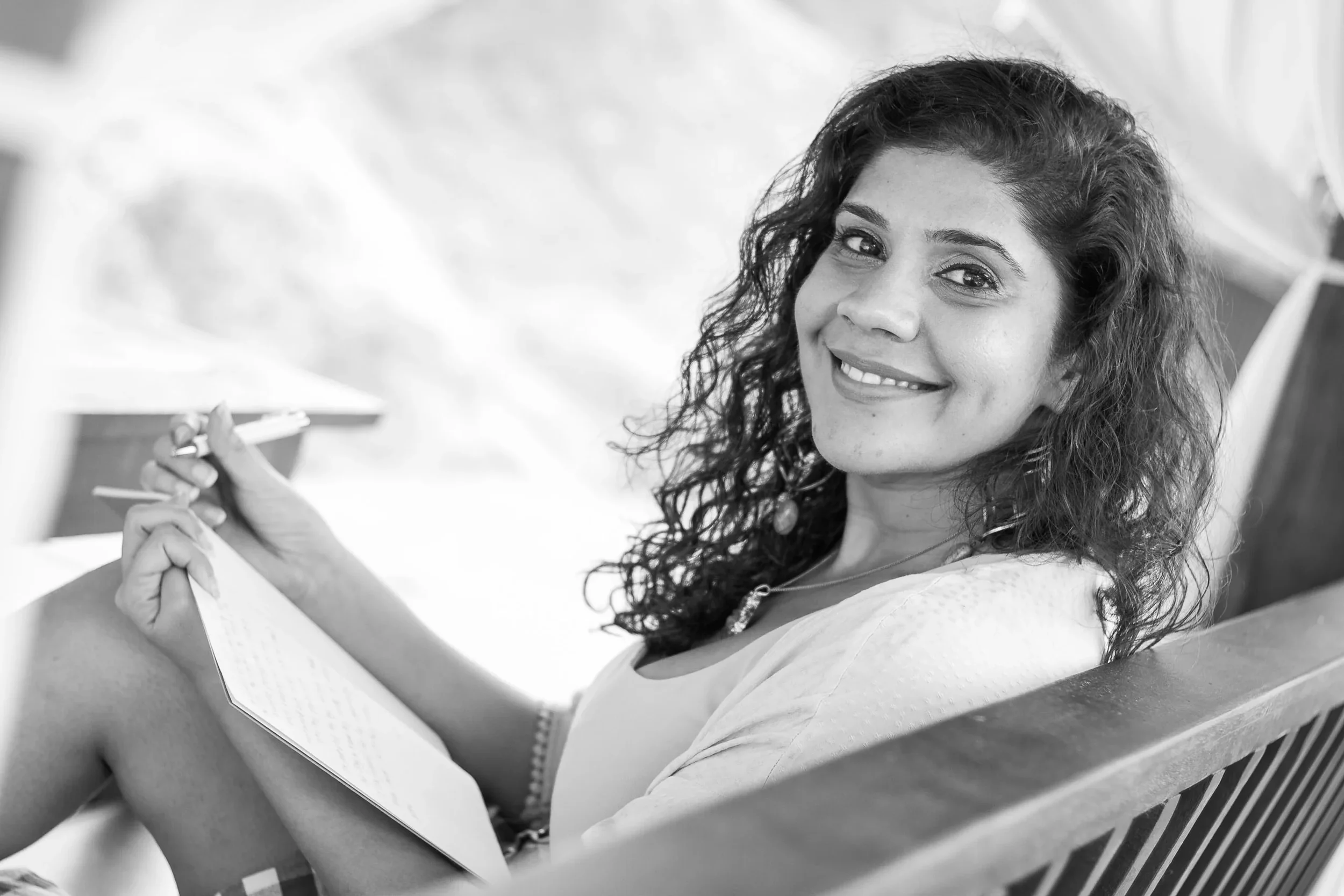 Black-and-white portrait of a curly-haired woman smiling warmly at the camera while holding a notebook and pen during a creative session, photographed by Seña Studio.
