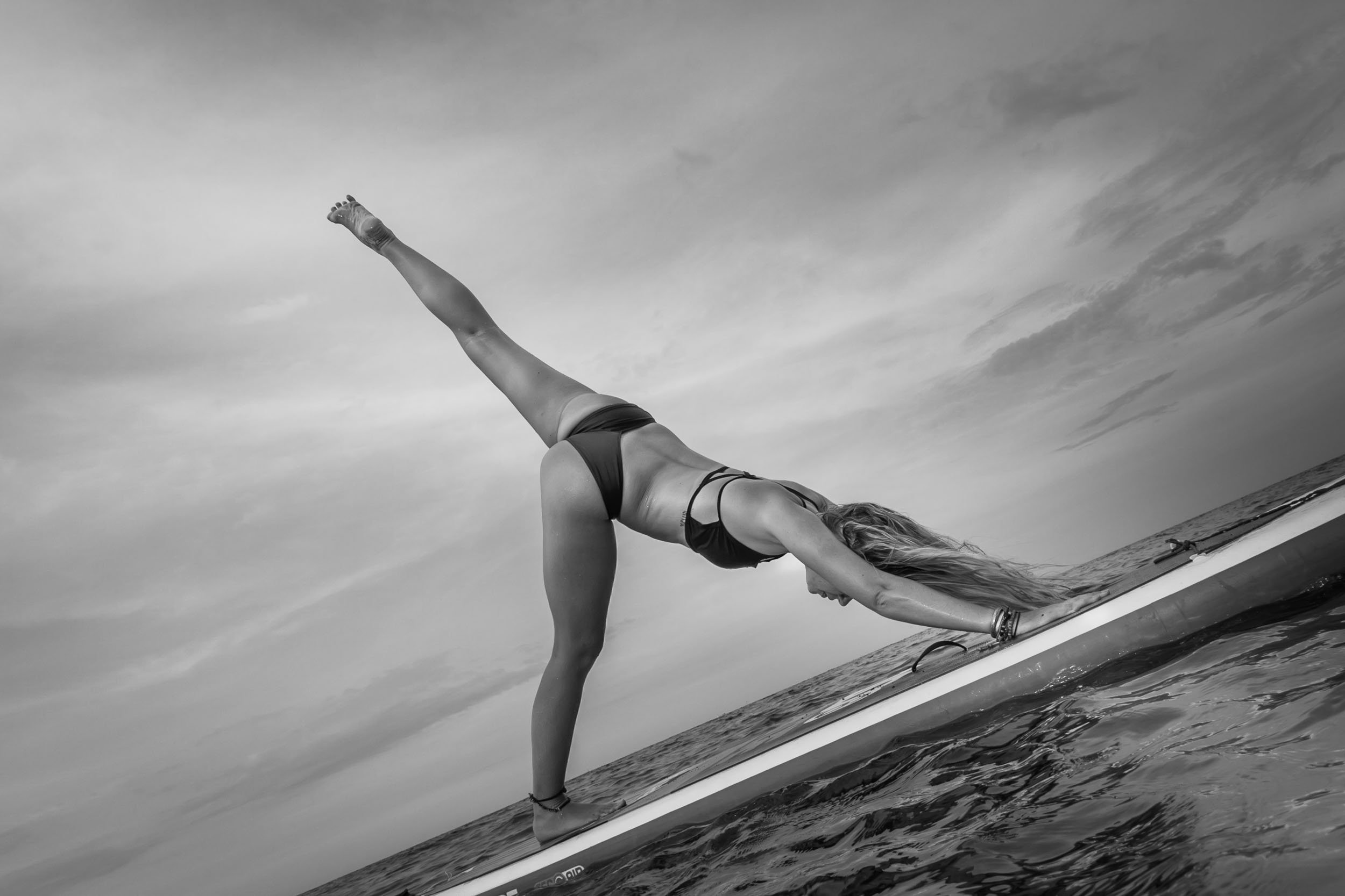 Dynamic low-angle black and white portrait capturing a woman balancing in a one-legged yoga extension on a paddleboard above calm water, photographed by Seña Studio.