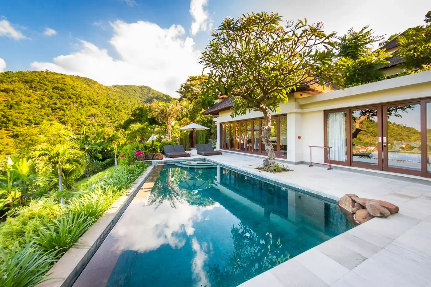 Private villa infinity pool reflecting clouds and surrounding mountains, complete with sun loungers and a frangipani tree at The Griya Villas & Spa in Amed, Bali, photographed by Seña Studio.