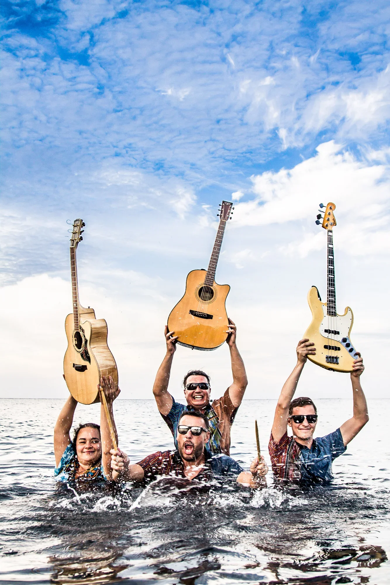 Dynamic portrait of a four-piece band led by Lucy Dobrovodska, wading waist-deep in ocean waves, cheering and holding their guitars and drumsticks high above the water, photographed by Seña Studio.