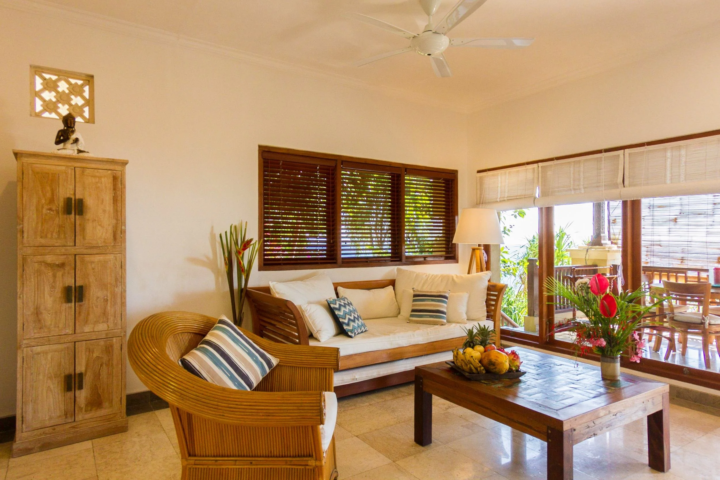 Warm, sunlit villa living room with wooden blinds, a woven rattan armchair, daybed couch, and tropical fruit platter on the coffee table at Aquamarine Beach Villas in Amed, Bali, photographed by Seña Studio.