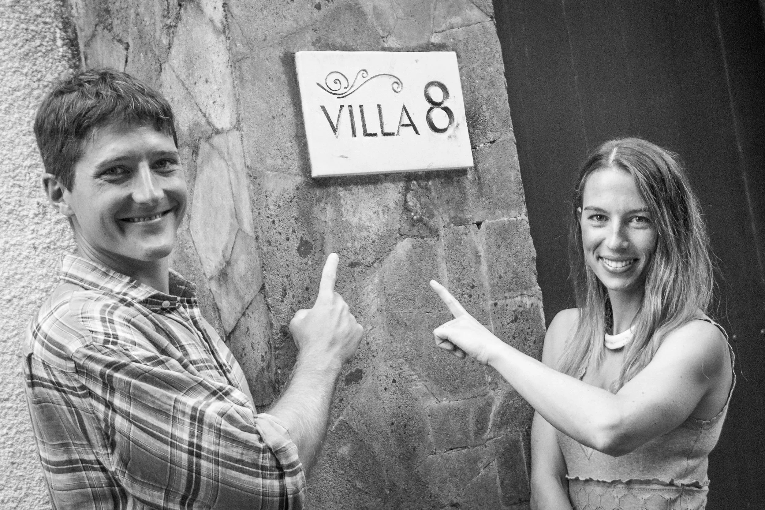 Playful black and white portrait of a smiling couple pointing up at a stone Villa 8 sign outside the entryway at The Griya Villas & Spa in Amed, Bali, photographed by Seña Studio.