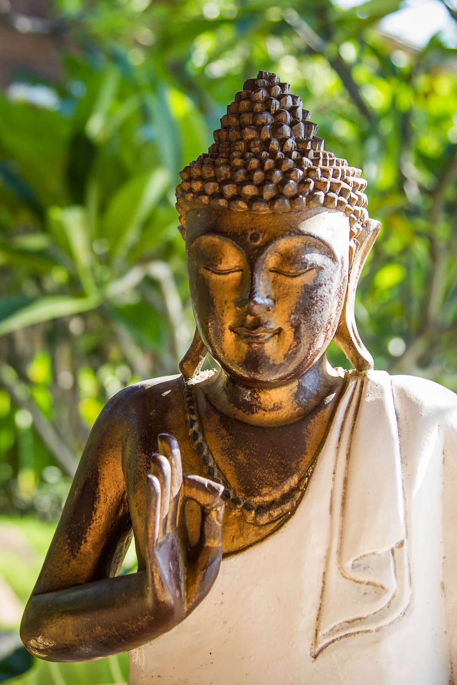 Warmly lit golden Buddha statue draped in a white cloth with eyes closed in a meditation pose, surrounded by blurred green resort gardens, photographed by Seña Studio.