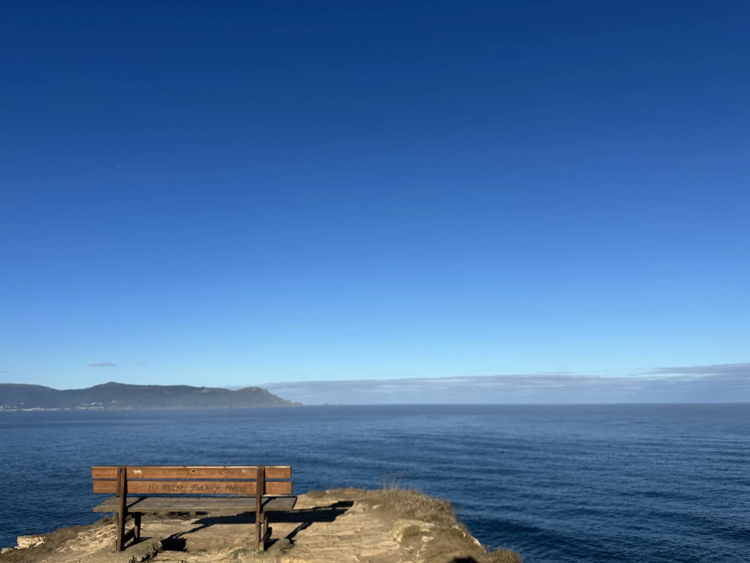 Wooden bench sitting high on a grassy cliff edge overlooking the deep blue Atlantic Ocean and coastline at Loiba in Northern Galicia, Spain, photographed by Seña Studio.