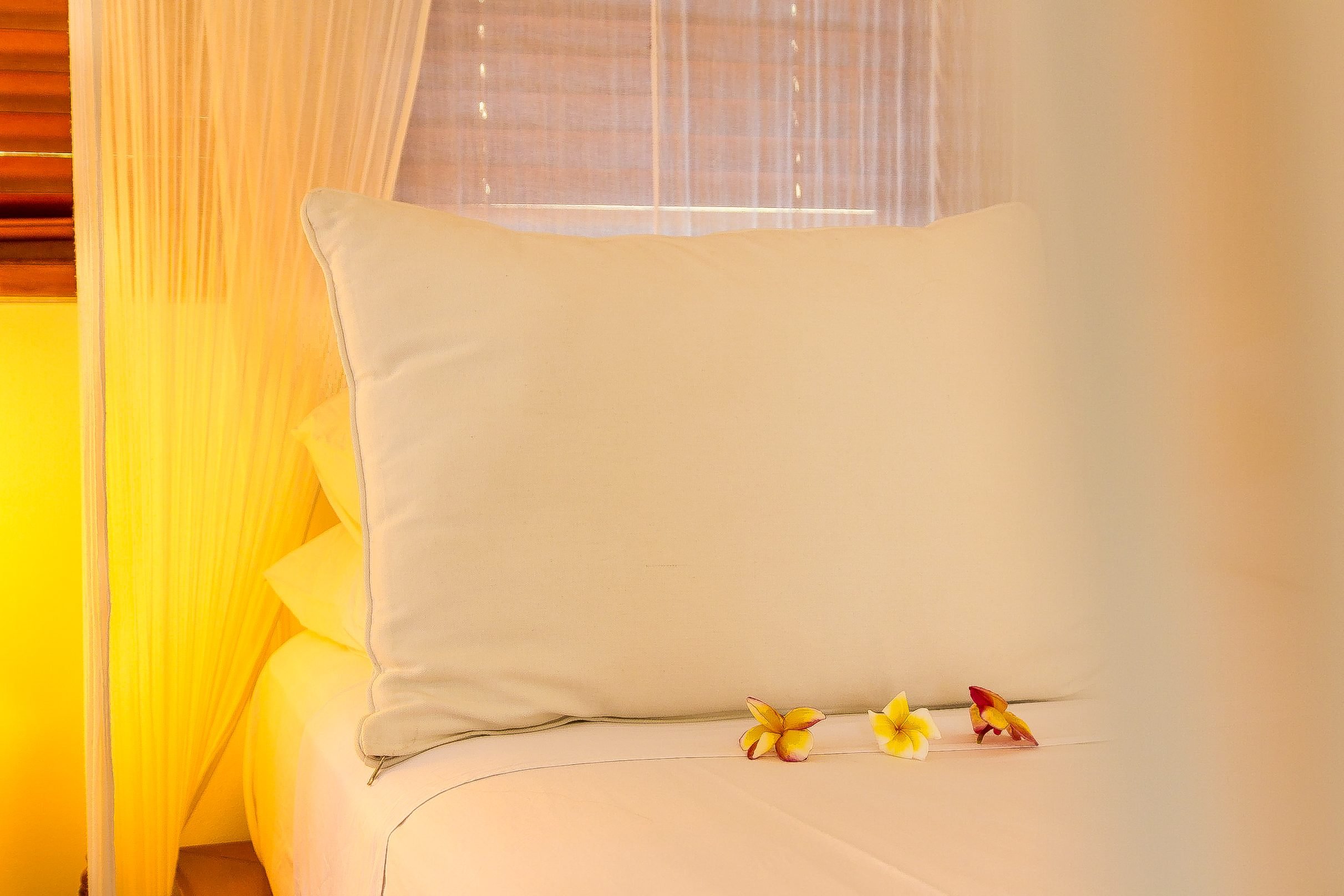 Close-up of crisp white bedding accented with fresh yellow and orange frangipani flowers in soft warm lighting at Aquamarine Beach Villas in Amed, Bali, photographed by Seña Studio.