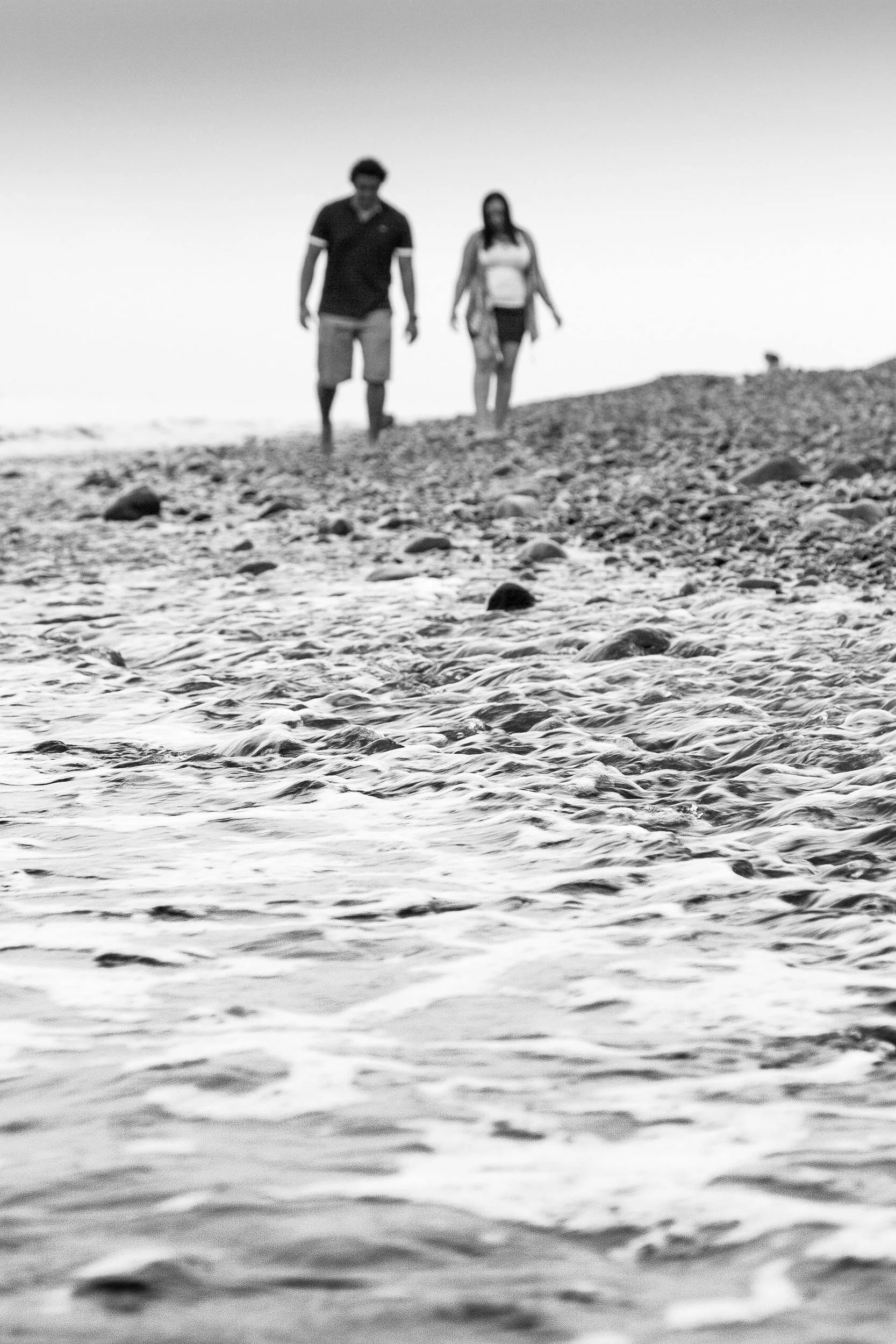Low-angle black-and-white artistic portrait focusing on water ripples on a pebble shore with a blurred couple walking together in the background, photographed by Seña Studio.