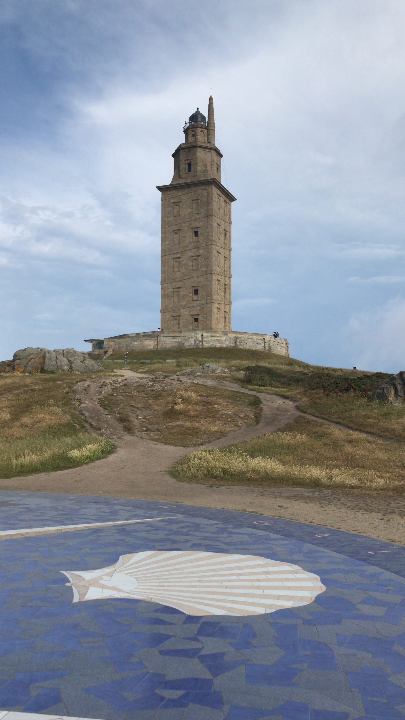 Vertical shot of the ancient Roman Tower of Hercules lighthouse rising above grassy hills with a blue scallop shell compass rose mosaic in the foreground in A Coruña, Galicia, photographed by Seña Studio.