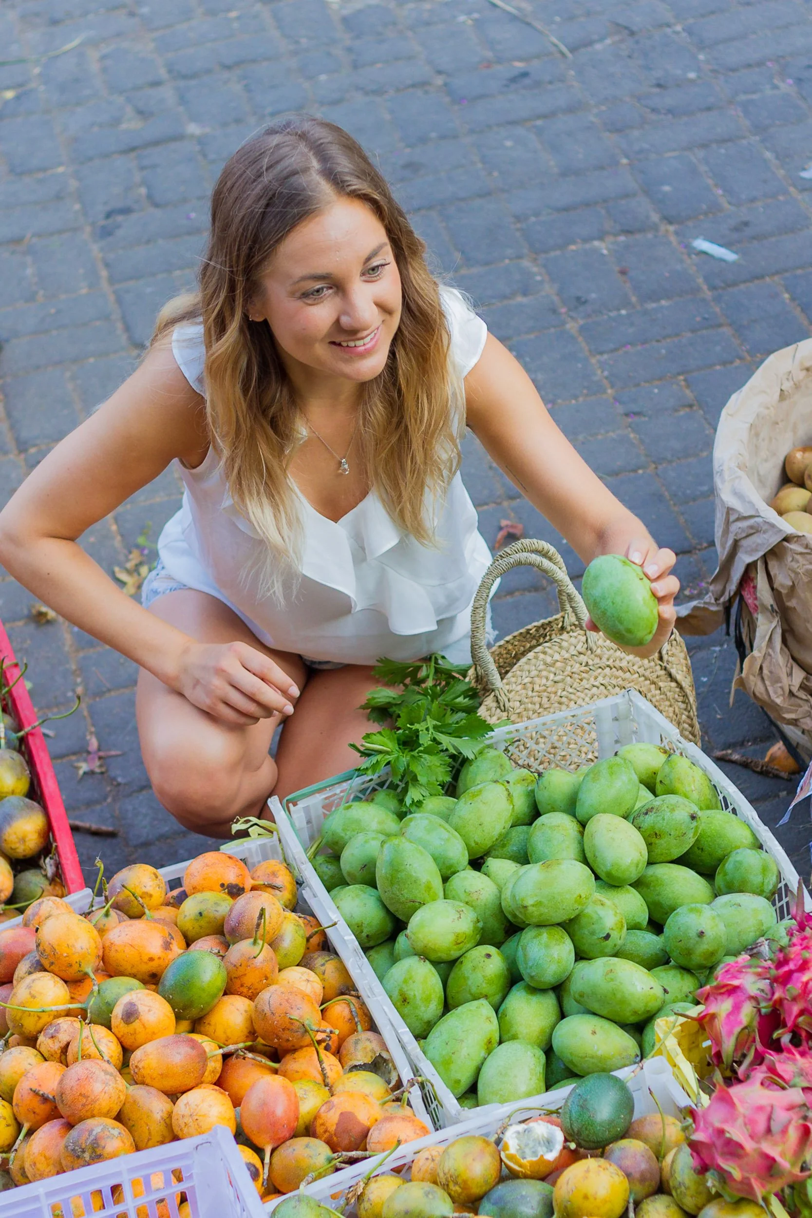 A blonde woman crouching down to pick out a green mango from a crate of fresh tropical fruits at an open-air market. Ubud, Bali.