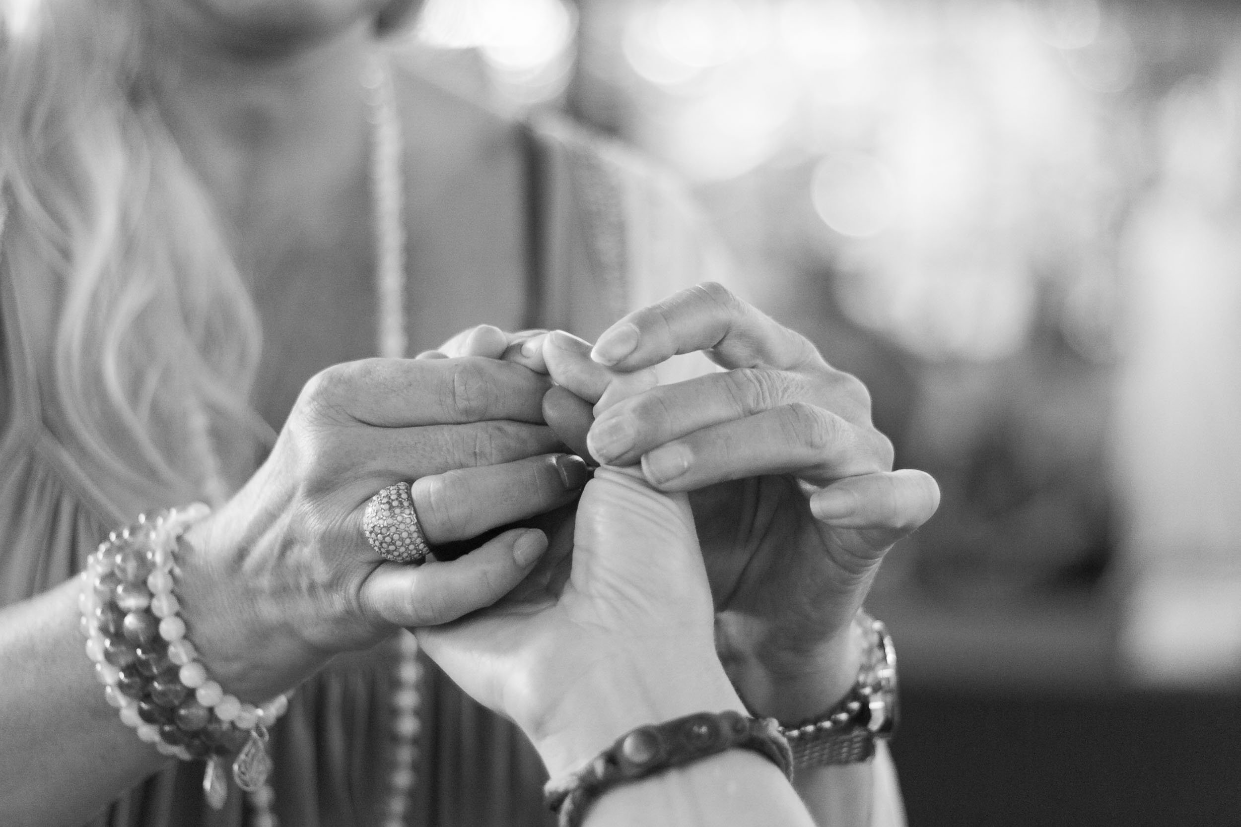 Soft black and white detail portrait showing a practitioner wearing beaded bracelets gently cradling a client's hands during a wellness session, photographed by Seña Studio.