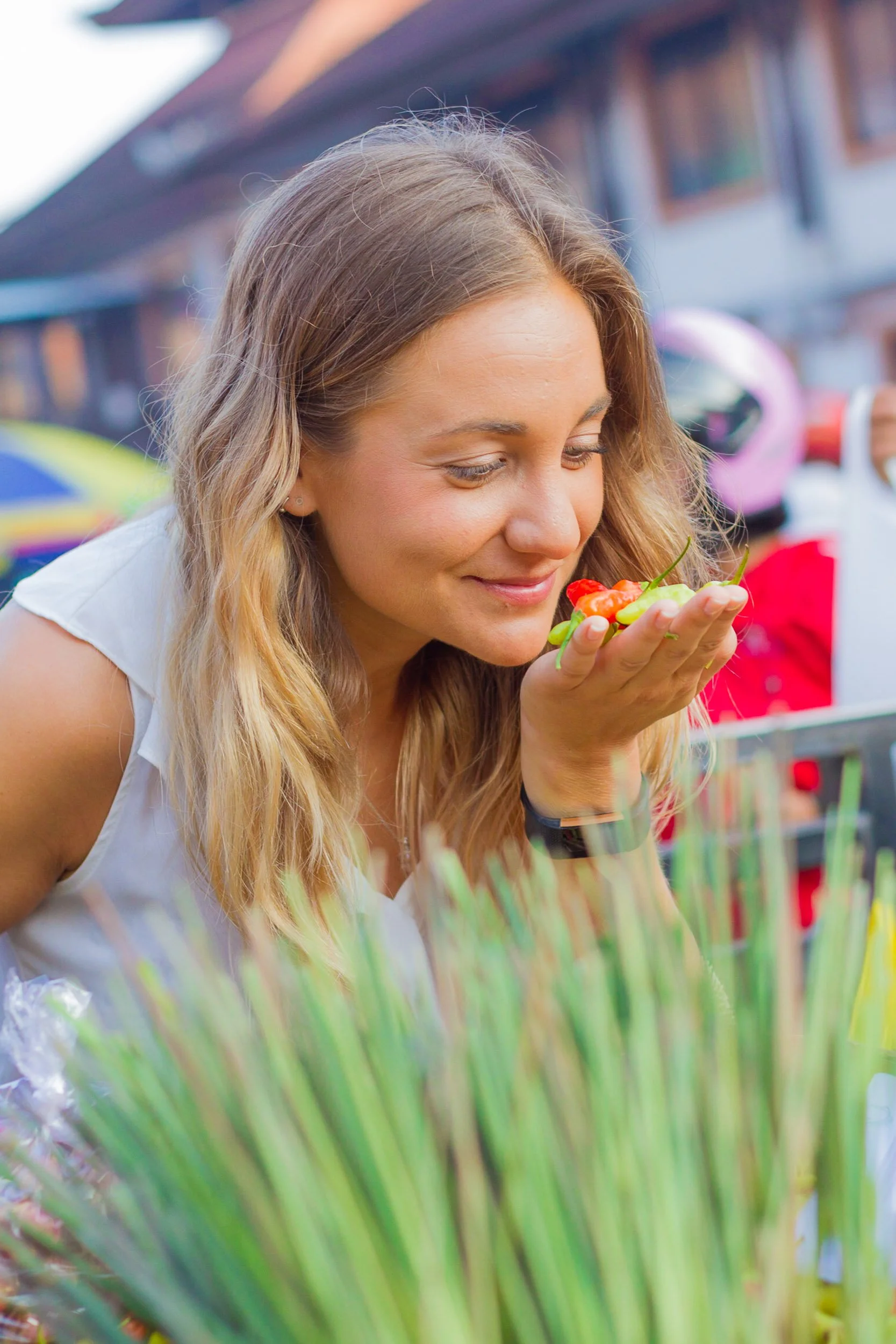 A smiling female chef smelling small fresh chili peppers held in her hand at a vibrant outdoor fresh produce market.