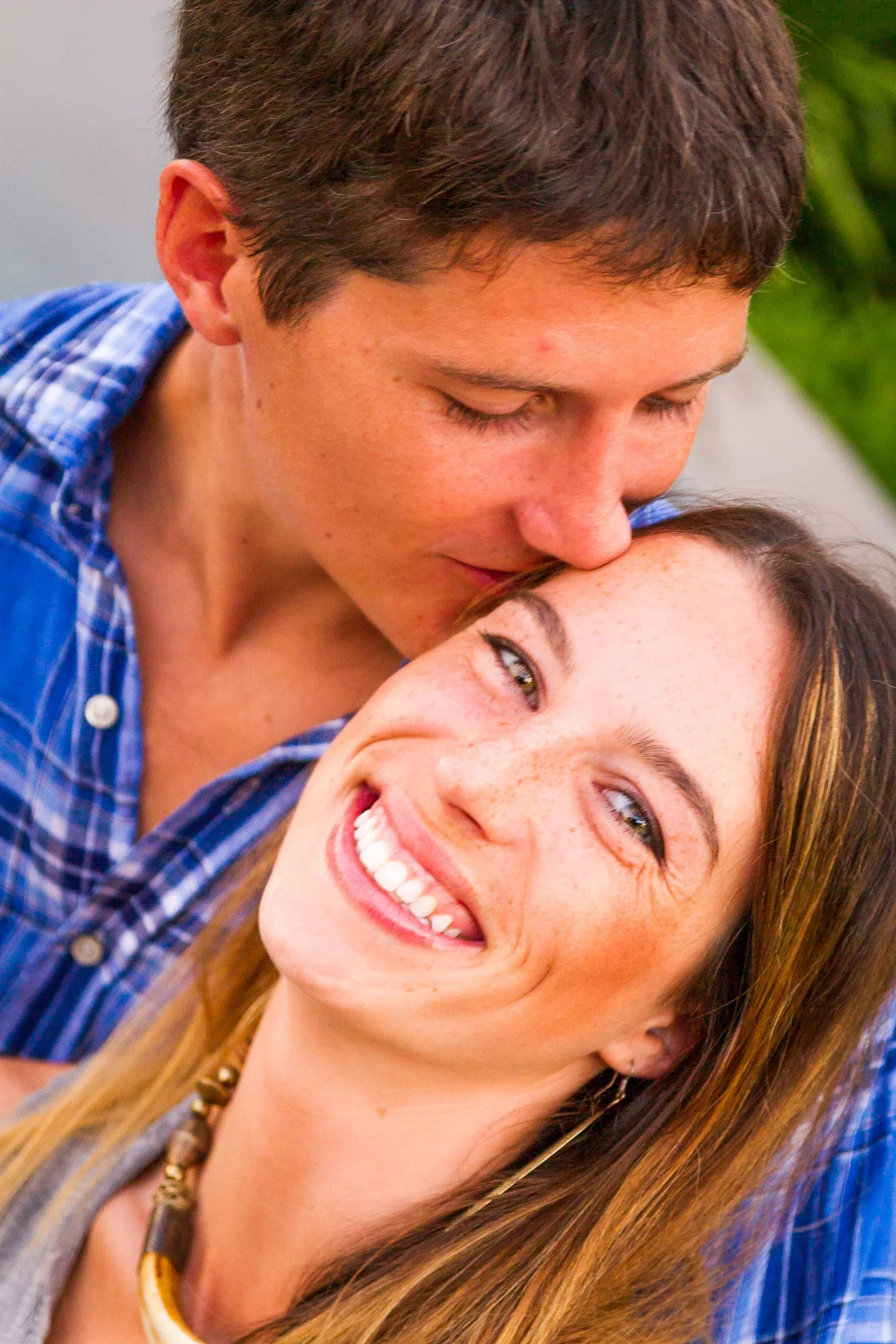Close-up natural light portrait of a man tenderly kissing a joyful, freckled woman on the forehead while she laughs toward the camera, photographed by Seña Studio.