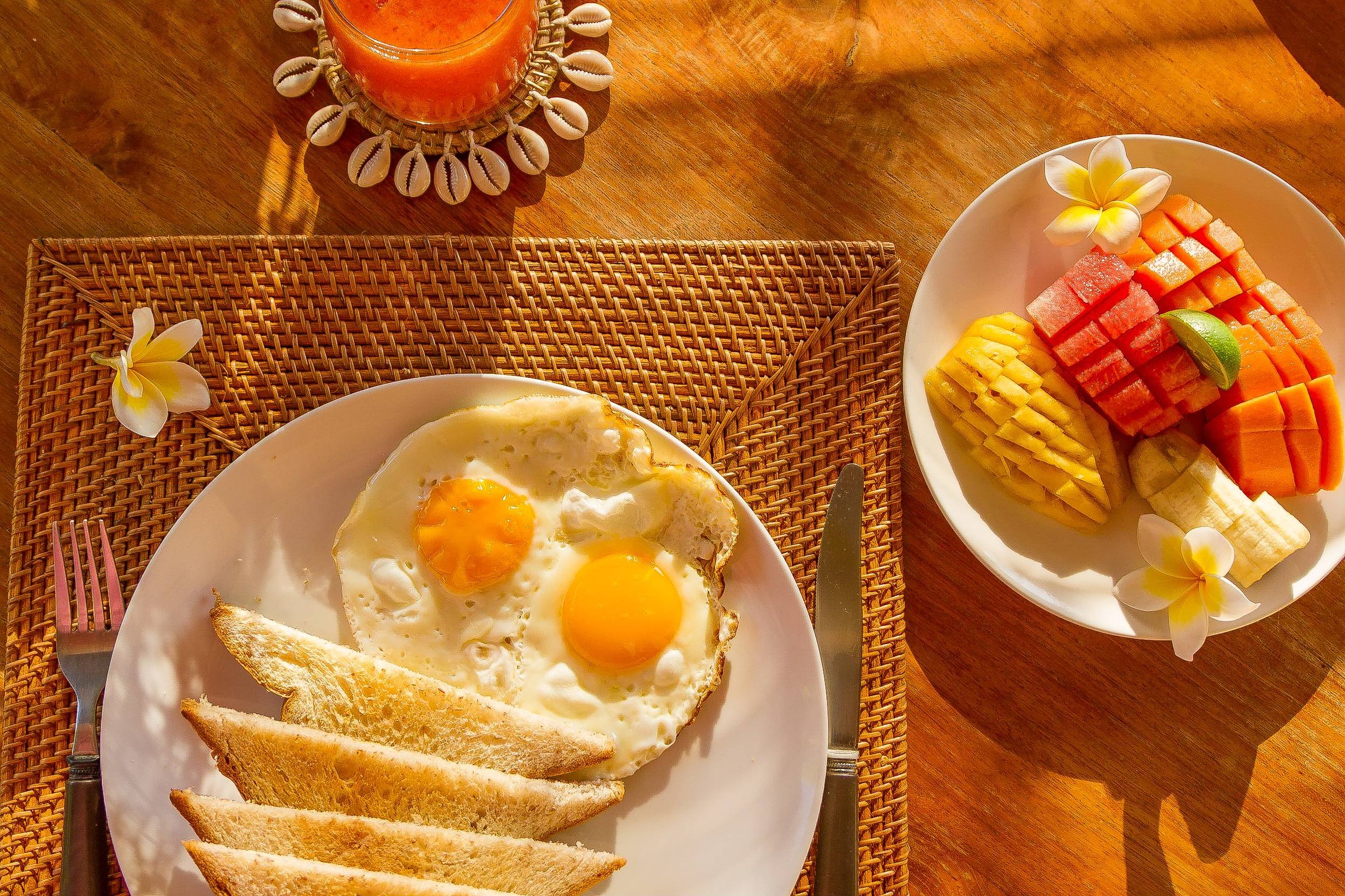 Overhead shot of a sunlit tropical breakfast set on a woven placemat, featuring fried eggs, toast, sliced papaya, pineapple, watermelon, and fresh juice garnished with frangipani blossoms, photographed by Seña Studio.