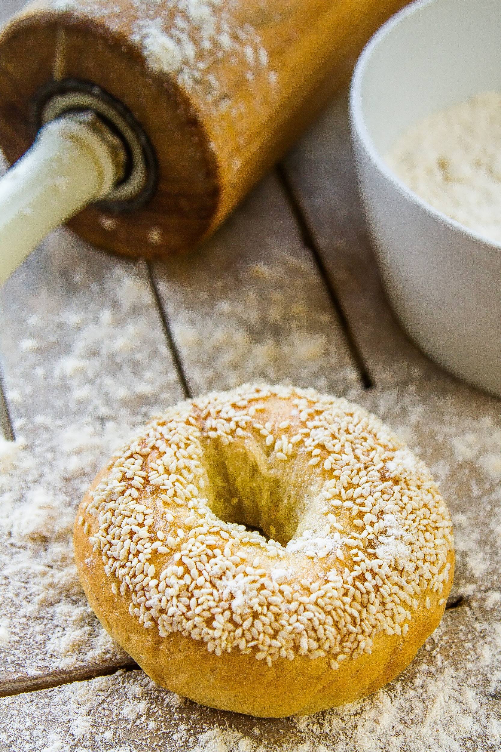 Vertical close-up of a golden freshly baked sesame seed bagel on a flour-dusted wooden prep counter next to a vintage rolling pin and bowl of flour, photographed by Seña Studio.
