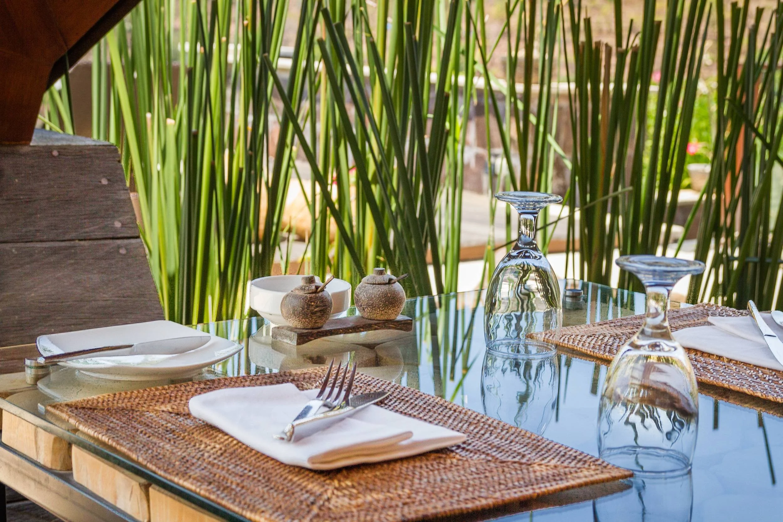 Close-up of an elegant resort restaurant table setting with woven placemats, folded white napkins, polished silverware, inverted water goblets, and wooden spice pots against green bamboo plants, photographed by Seña Studio.