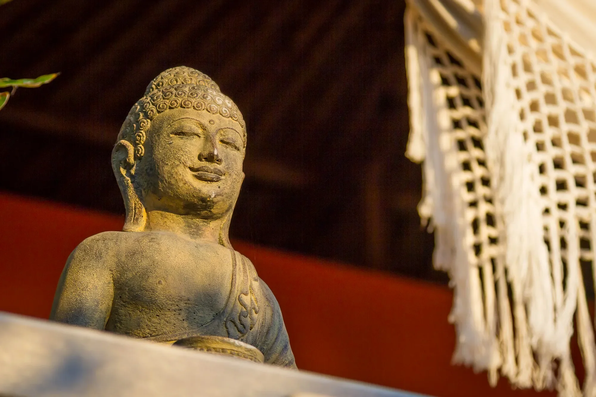 Serene stone Buddha statue illuminated by warm sunlight next to fringe macramé decor at Aquamarine Beach Villas in Amed, Bali, photographed by Seña Studio.