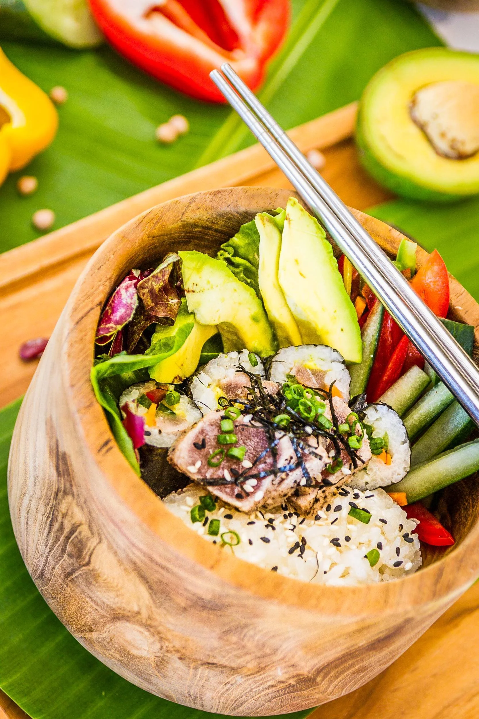 Wooden bowl filled with seared tuna slices, sushi rolls, avocado slices, cucumber, and sesame seeds with metal chopsticks rested on top, photographed by Seña Studio.