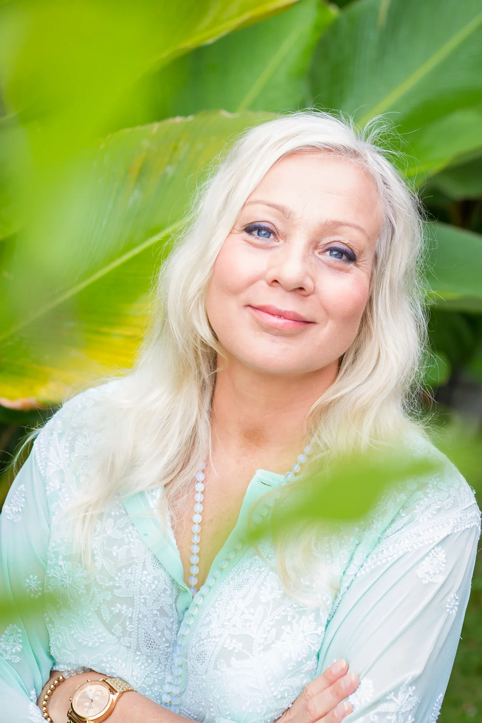 Portrait of a blonde woman with blue eyes wearing a mint green embroidered blouse with vibrant green tropical leaves in the background.