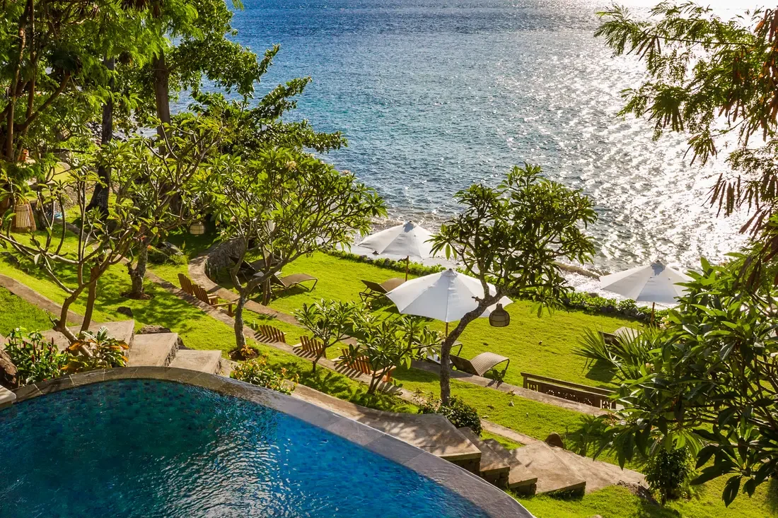 Terraced oceanfront lawn at Aquamarine Beach Villas in Amed, Bali, with white sun umbrellas, lounge chairs, and palm trees, overlooking the shimmering sea, photographed by Seña Studio.
