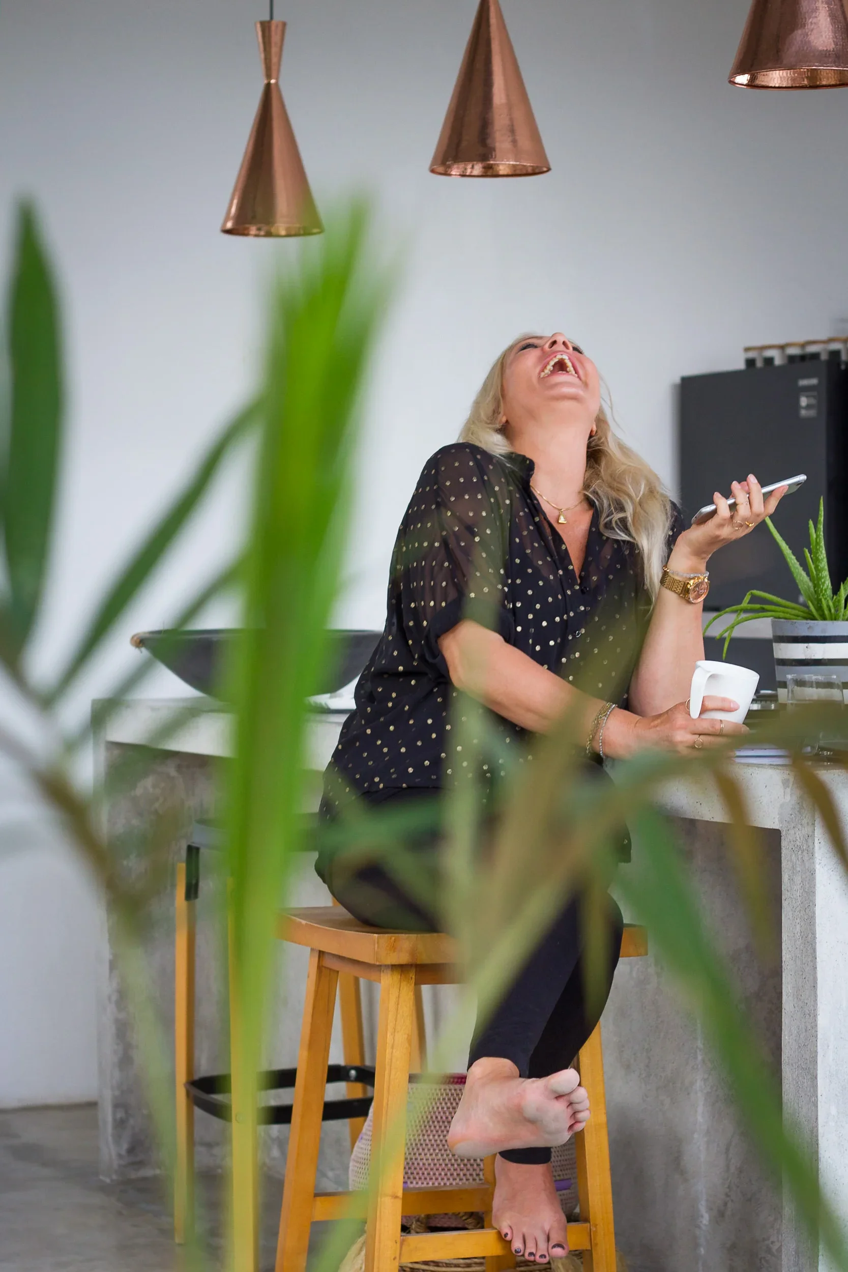Blonde woman holding a coffee cup sitting on a high stool laughing with her head tilted back.