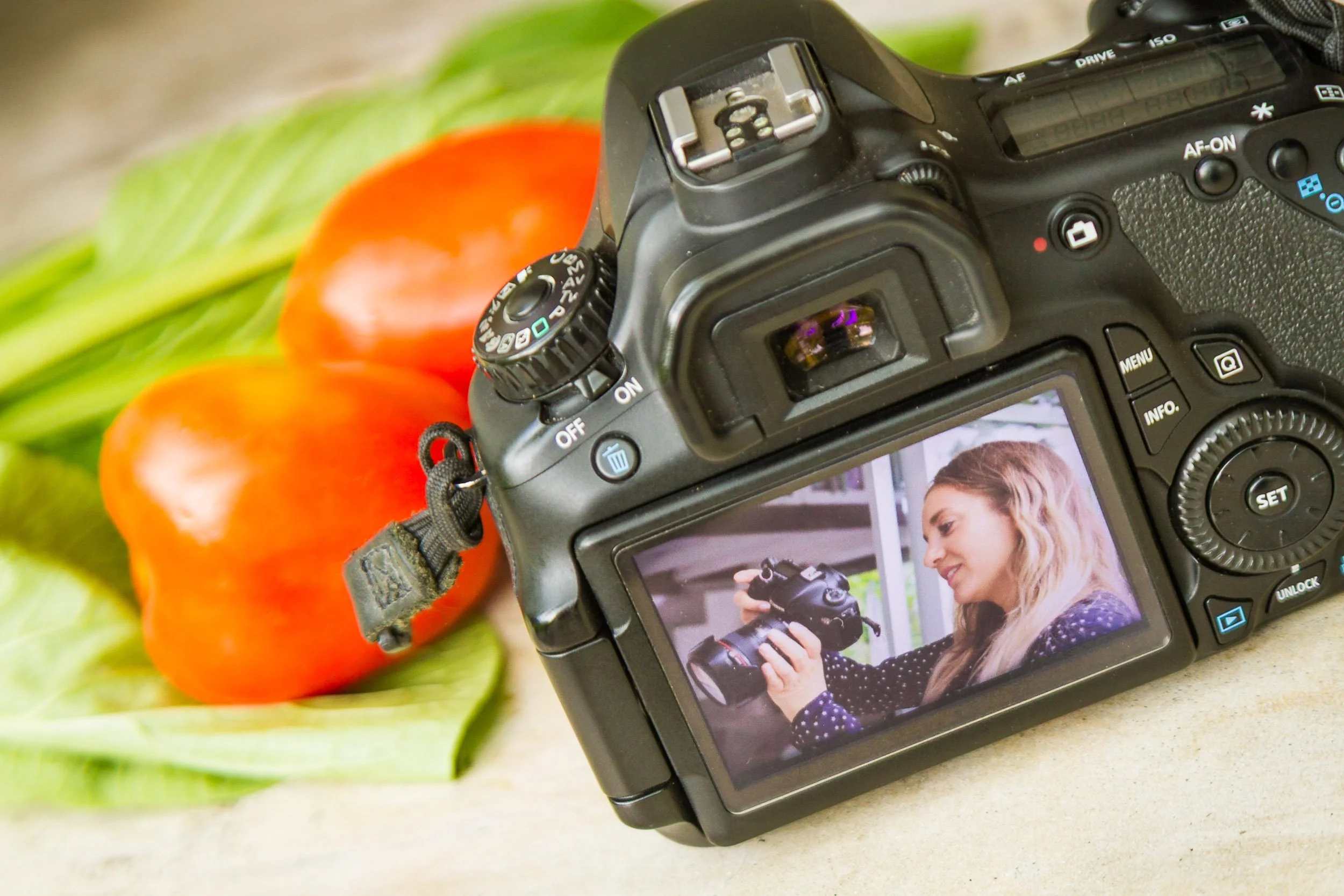 A close-up view of a professional DSLR camera screen displaying a female photographer, set beside fresh red bell peppers and leafy green vegetables. Behind-the-scenes shot.