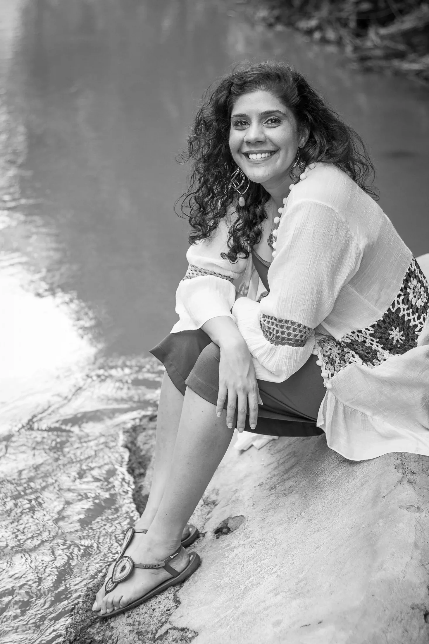 Black and white outdoor portrait of a laughing curly-haired woman in a embroidered bohemian blouse sitting along a natural riverbank, photographed by Seña Studio.