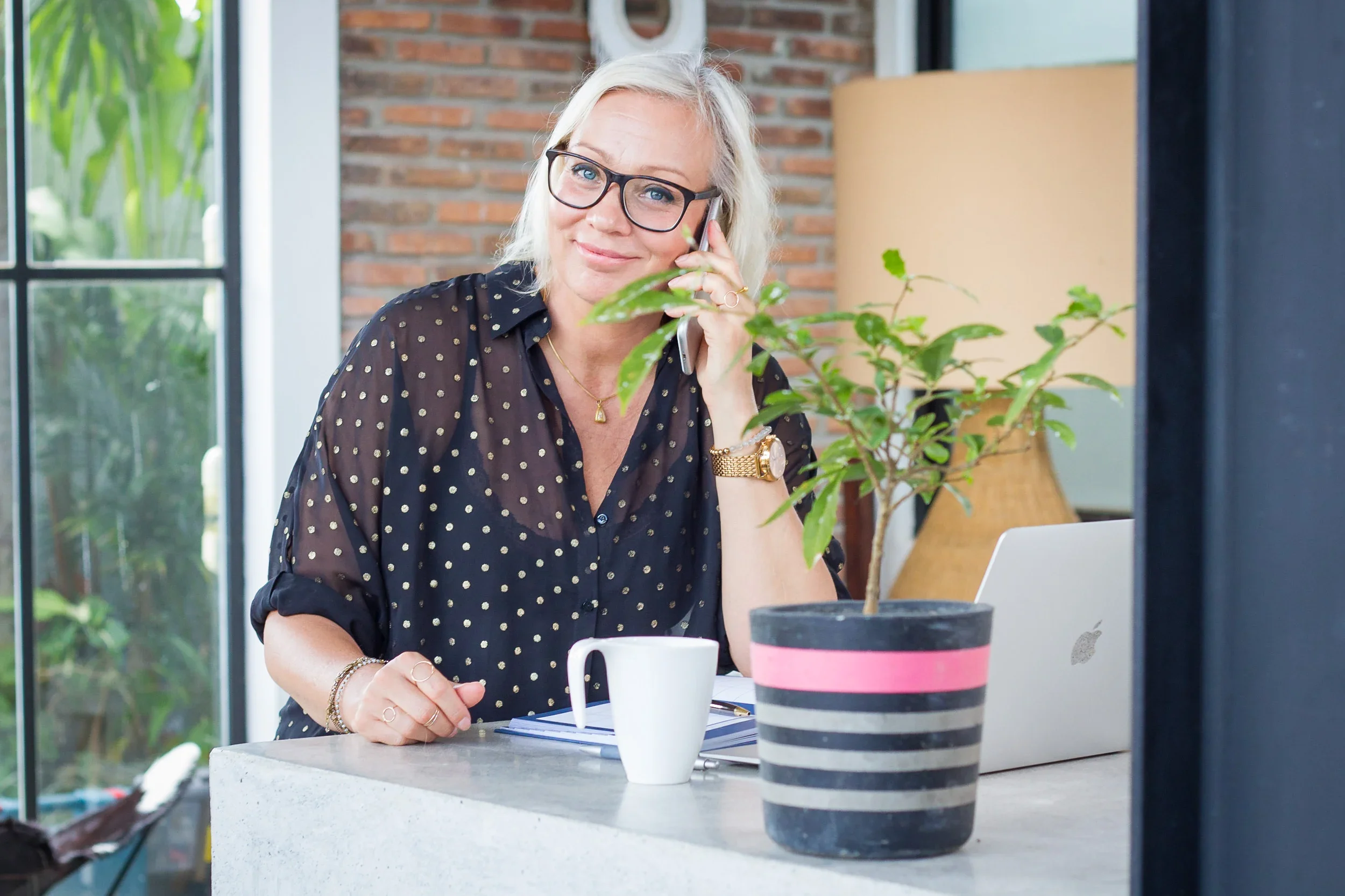 Blonde woman in black glasses and a gold polka dot shirt talking on the phone at a modern concrete counter desk.
