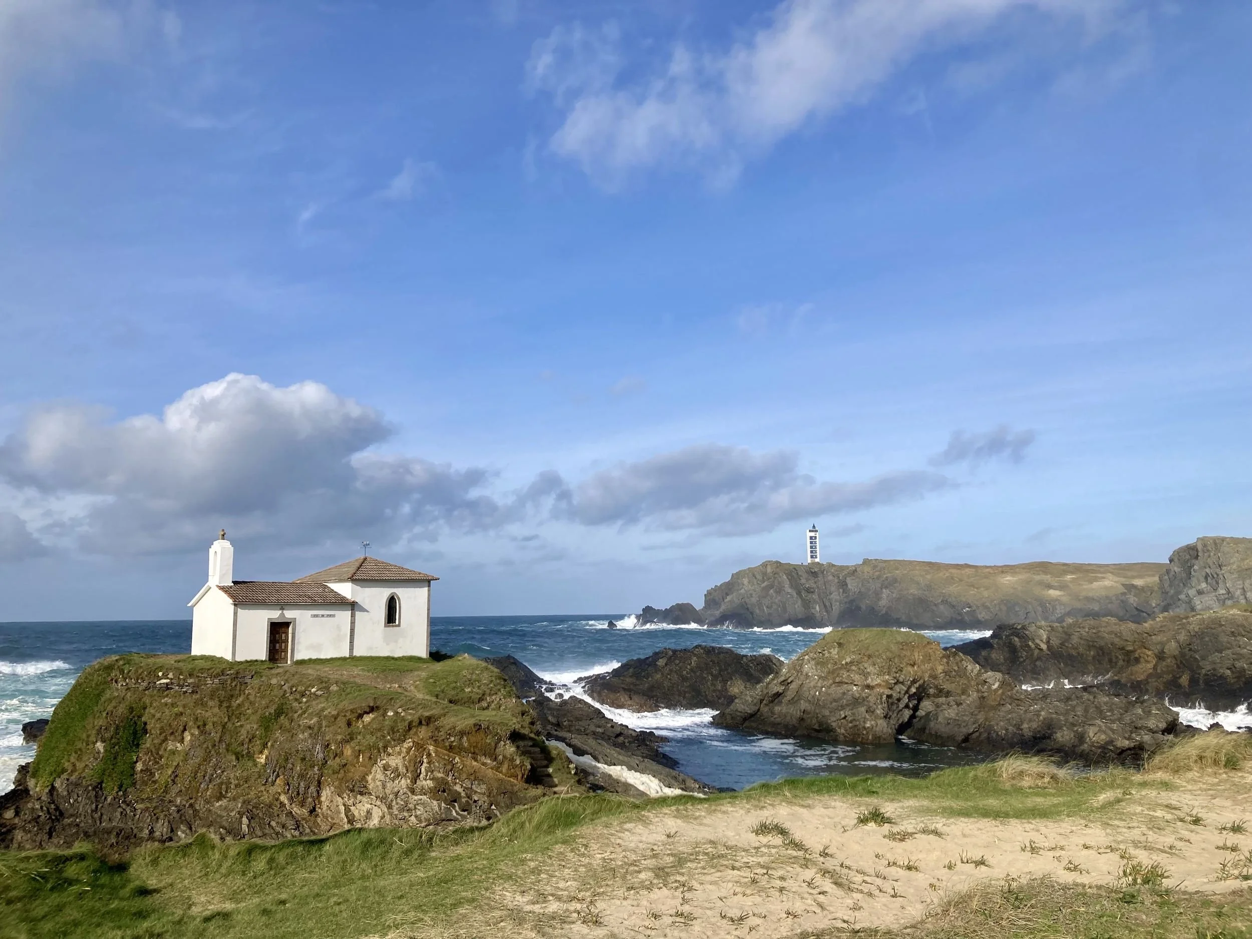 Historic white Ermita de la Virgen del Puerto chapel sitting atop a rocky coastal promontory with ocean waves breaking and a distant lighthouse in Galicia, photographed by Seña Studio.