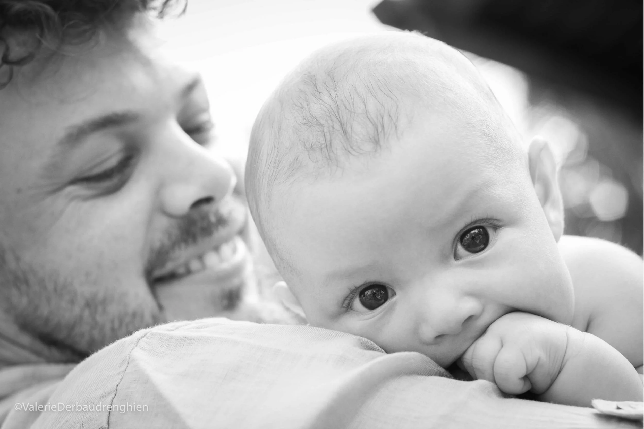 Baby peeks over dad’s shoulder with a fist in mouth while father smiles, a black and white family photo