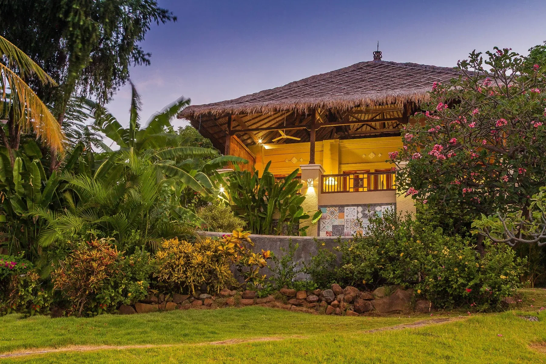 Exterior view of a luxury thatched-roof tropical villa warmly illuminated at dusk, surrounded by lush palm trees and flowering gardens at Aquamarine Beach Villas in Amed, Bali, photographed by Seña Studio.