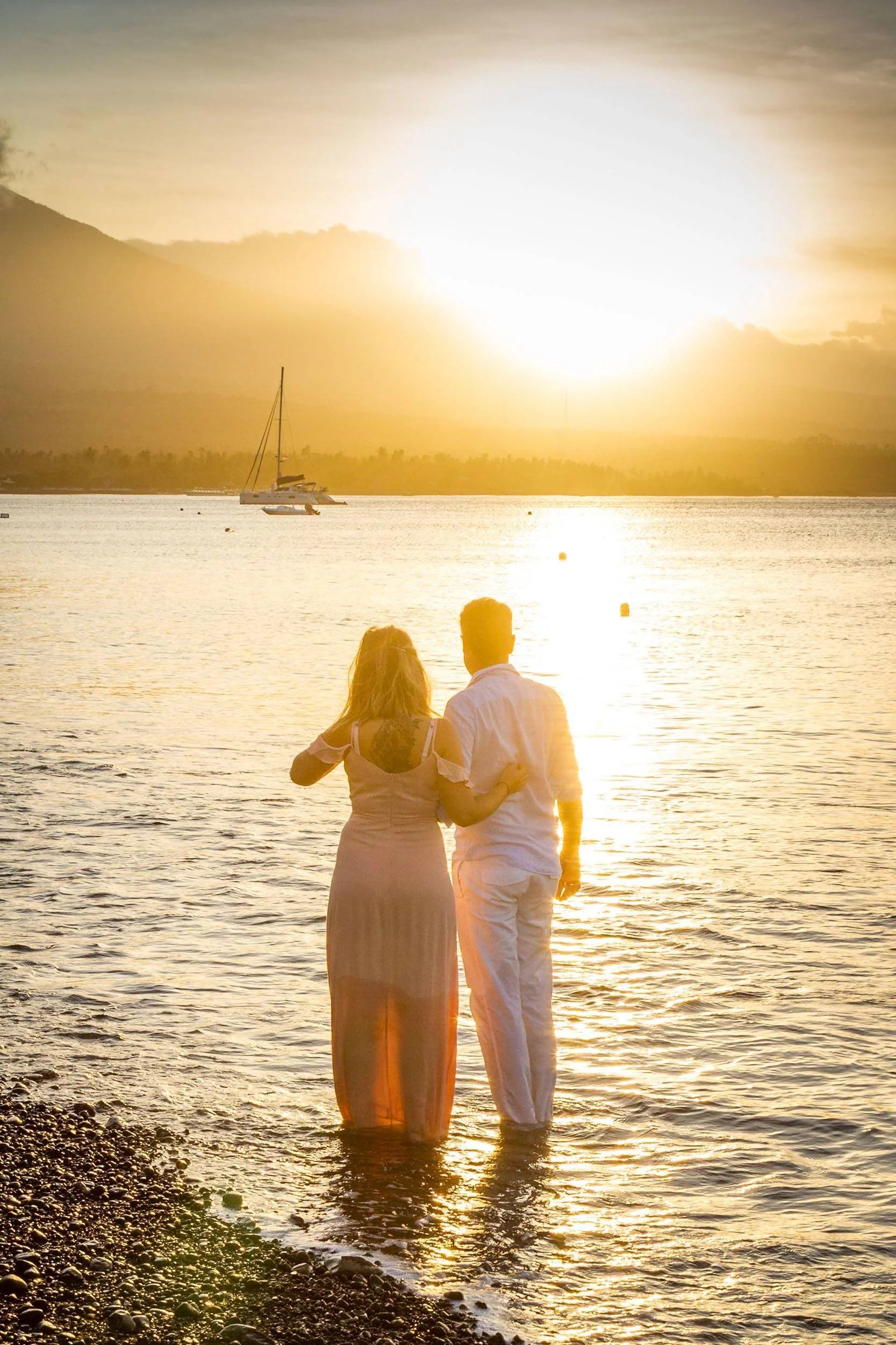 Back-view portrait of a couple standing ankle-deep in calm ocean water at sunset, embraced as a sailboat rests on the golden horizon, photographed by Seña Studio.