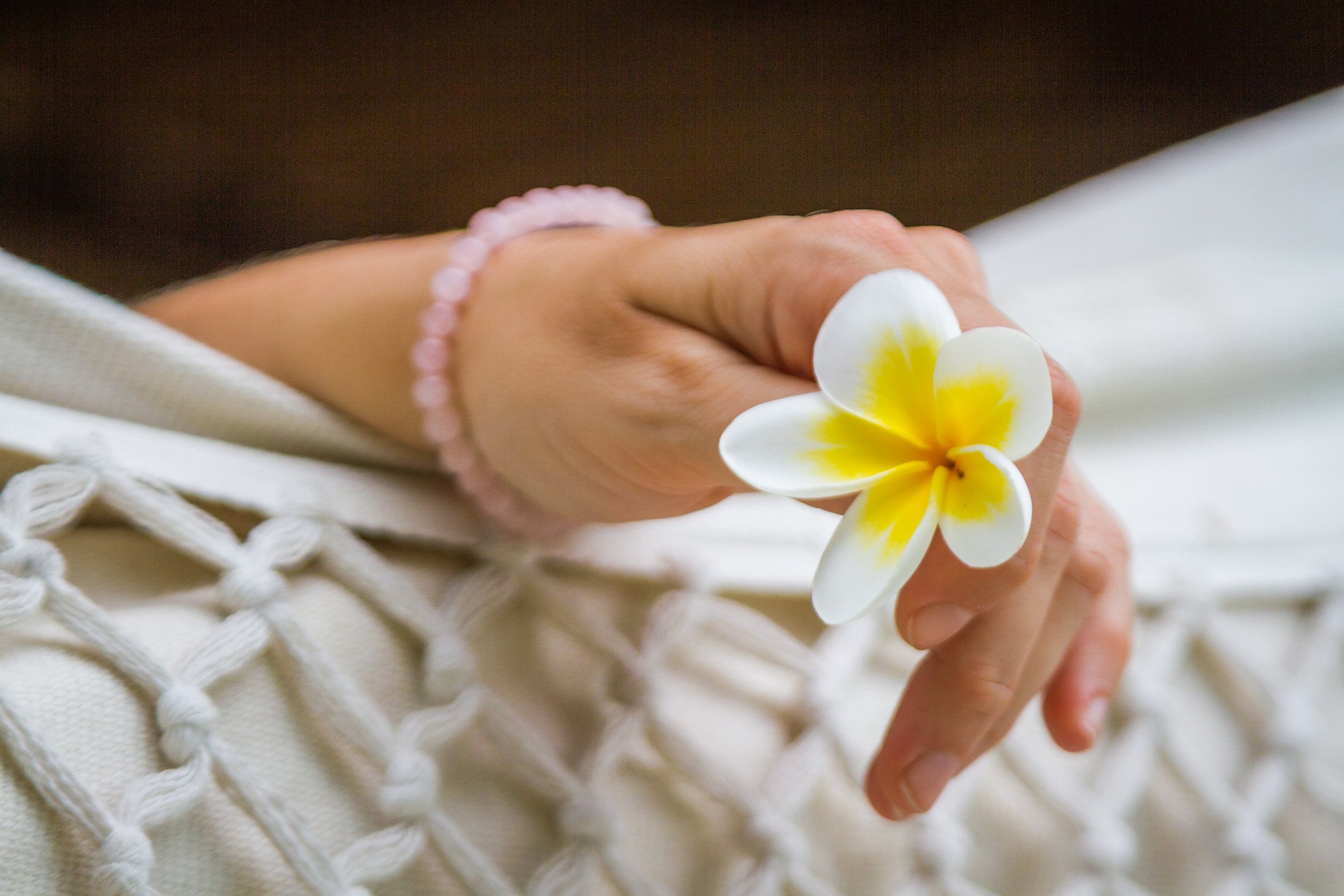 Detailed close-up of a woman wearing a rose quartz beaded bracelet holding a white and yellow frangipani flower while relaxing in a white macramé hammock, photographed by Seña Studio.